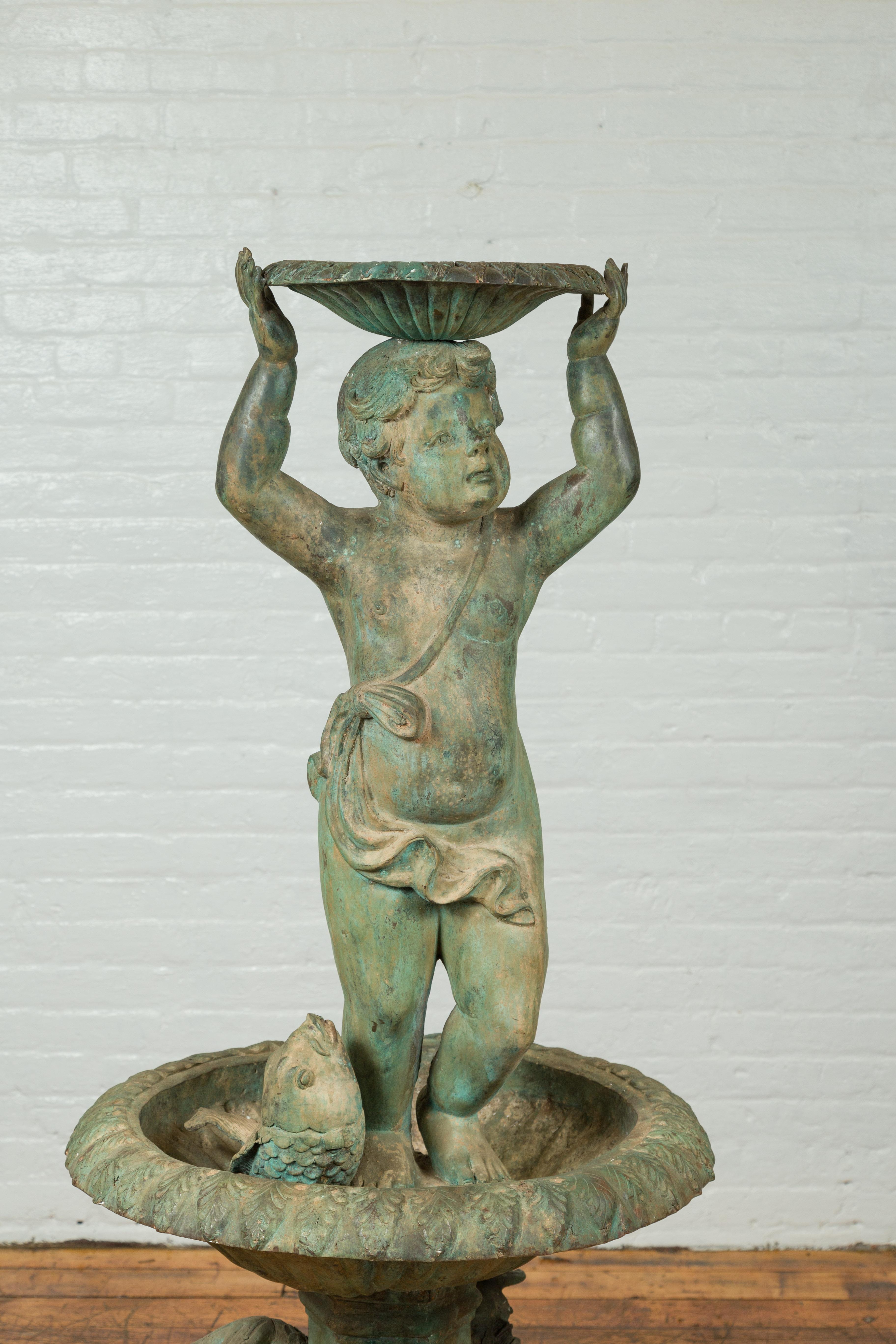A Greco Roman style vintage putto fountain with distressed verde patina, basin and fish motifs. Created with the traditional technique of the lost-wax (à la cire perdue) that allows a great precision in the details, this bronze fountain captures our