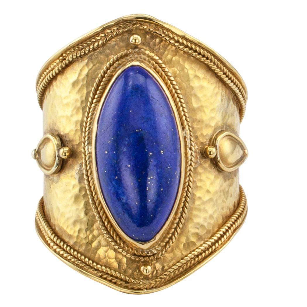 Greek 1990s lapis lazuli gold ring band.  The Etruscan style design features a bezel-set lapis lazuli on a wide and tapering 22-karat yellow gold ring band with a hammered texture and decorated with corded gold motifs, granulation and applied gold
