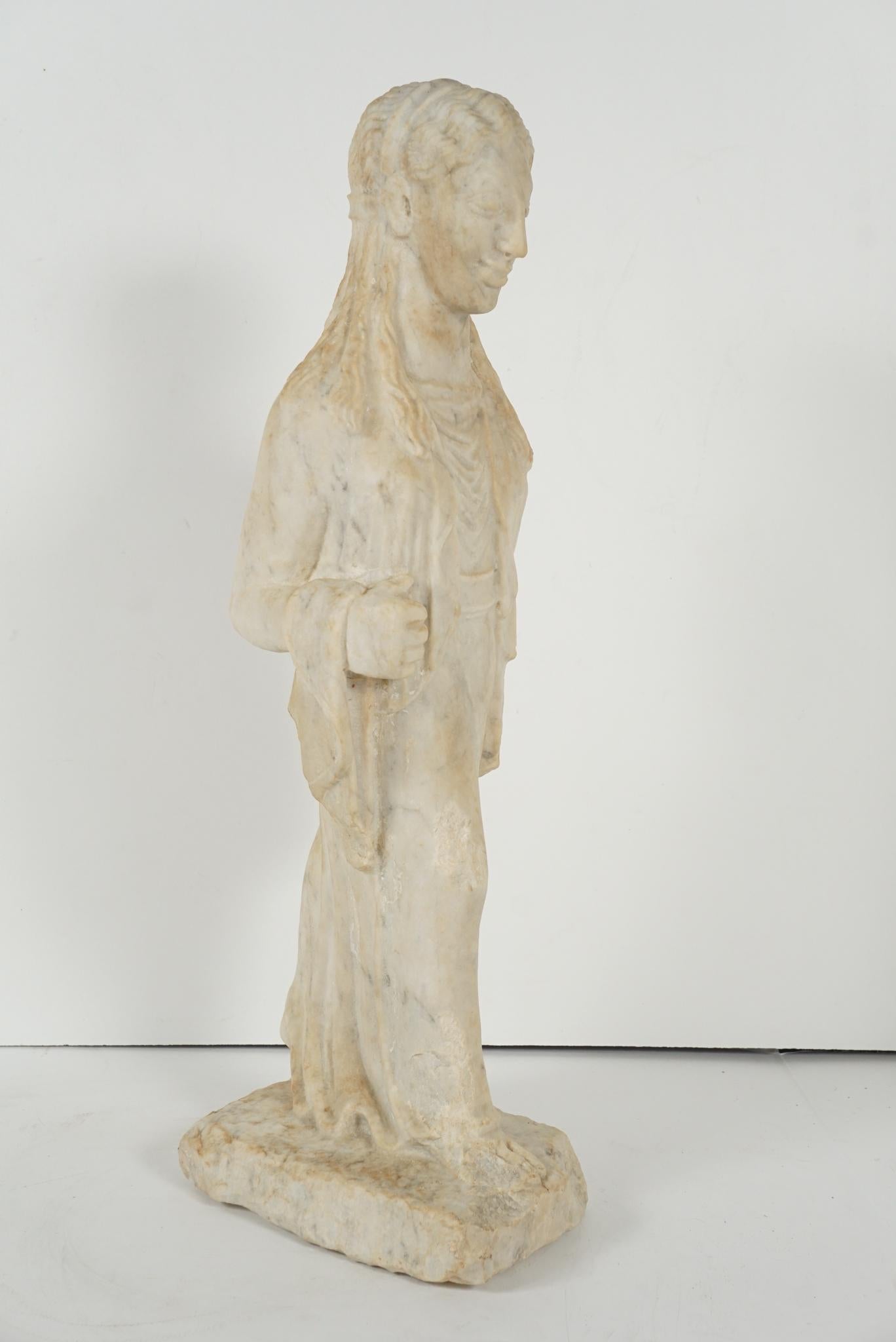 Created, circa 1910 this carved marble figure called a Kore is done in the Archaistic style of ancient Greece and was made as a Grand Tour item to return home with after a classical education abroad. The figure of a female is classically draped and