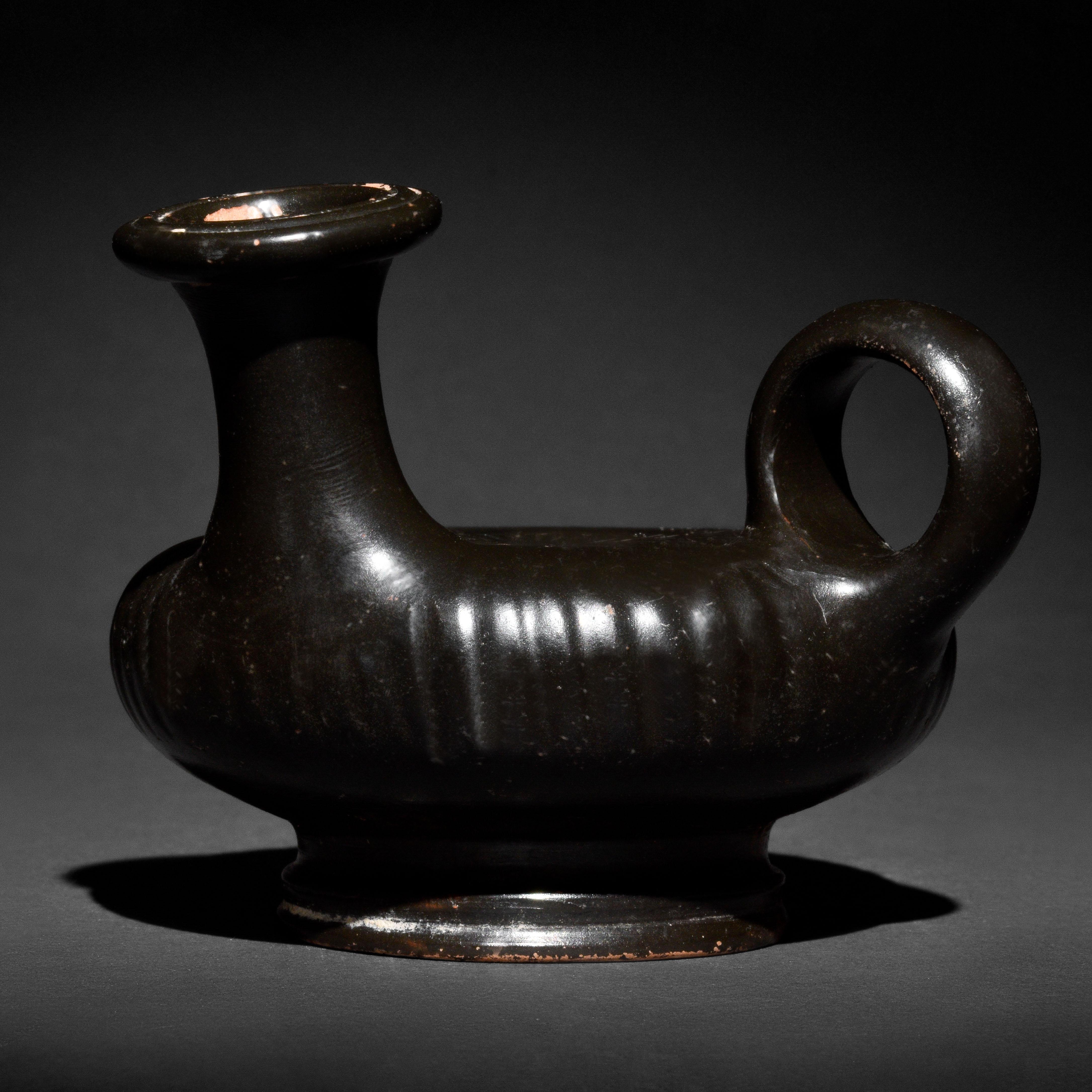 A black-glazed terracotta guttus (oil-lamp filler) with a spool-shaped foot, a wheel-shaped body, a protruding ring handle, and a cylindrical spout with an everted rim. The tondo features a rosette. This type of vessel, a guttus, was used to store