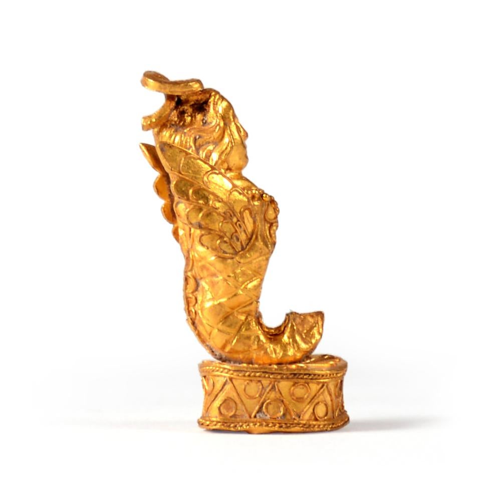A gold pendant in the shape of a siren with a human head, hair parted and falling onto back, human upper body, wings on back, standing on drum-shaped pedestal, decorated with granules and threading.

2,9 cm (h)

Ex European private collection, Mr.
