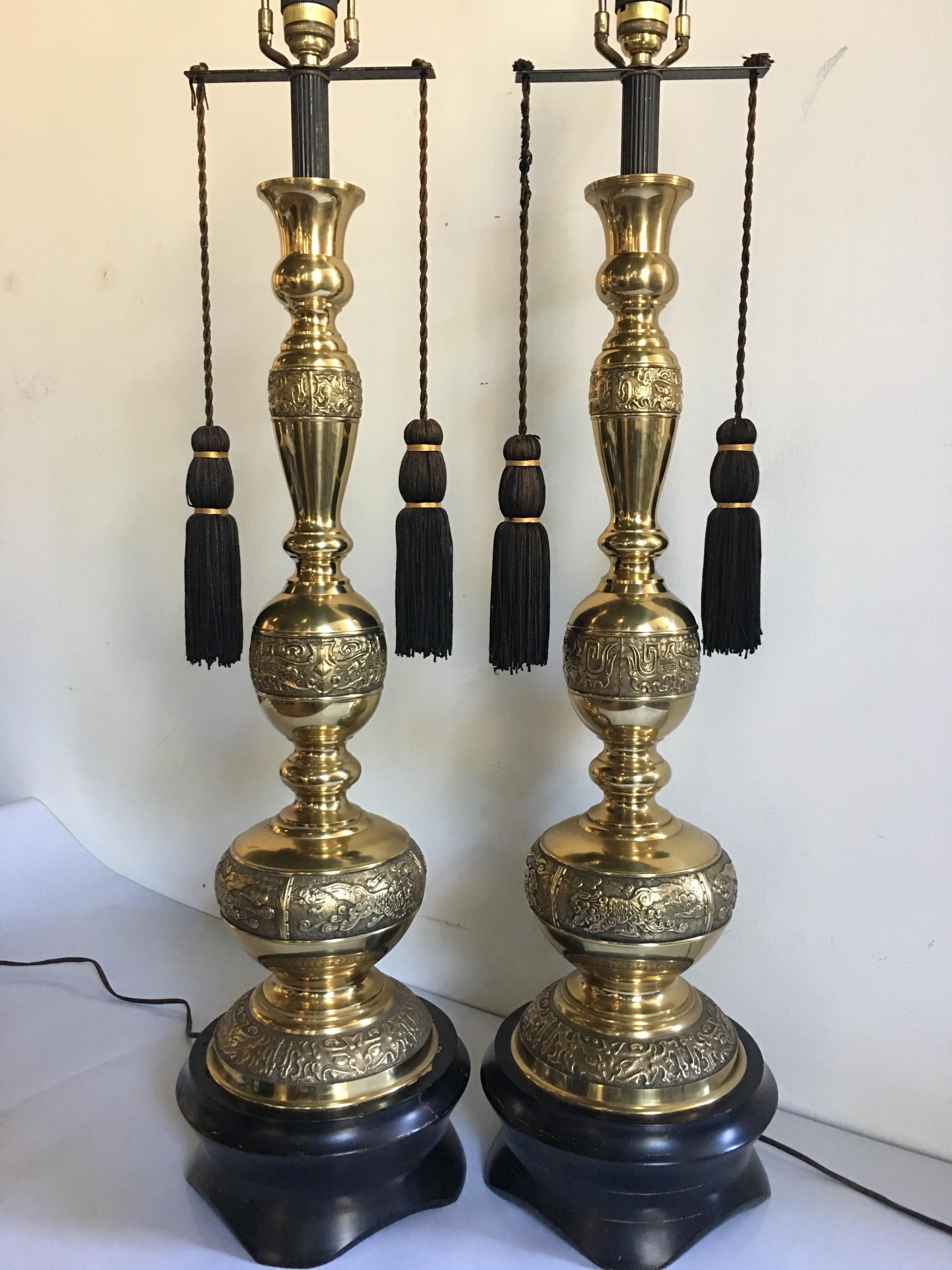 Pair of massive Hollywood Regency style finely cast brass lamps with Asian / Chinese and Greek key inspired motifs. These tall chinoiserie style table lamps features decorative tassels and are mounted on black plinth wood bases. Lamps turn on and