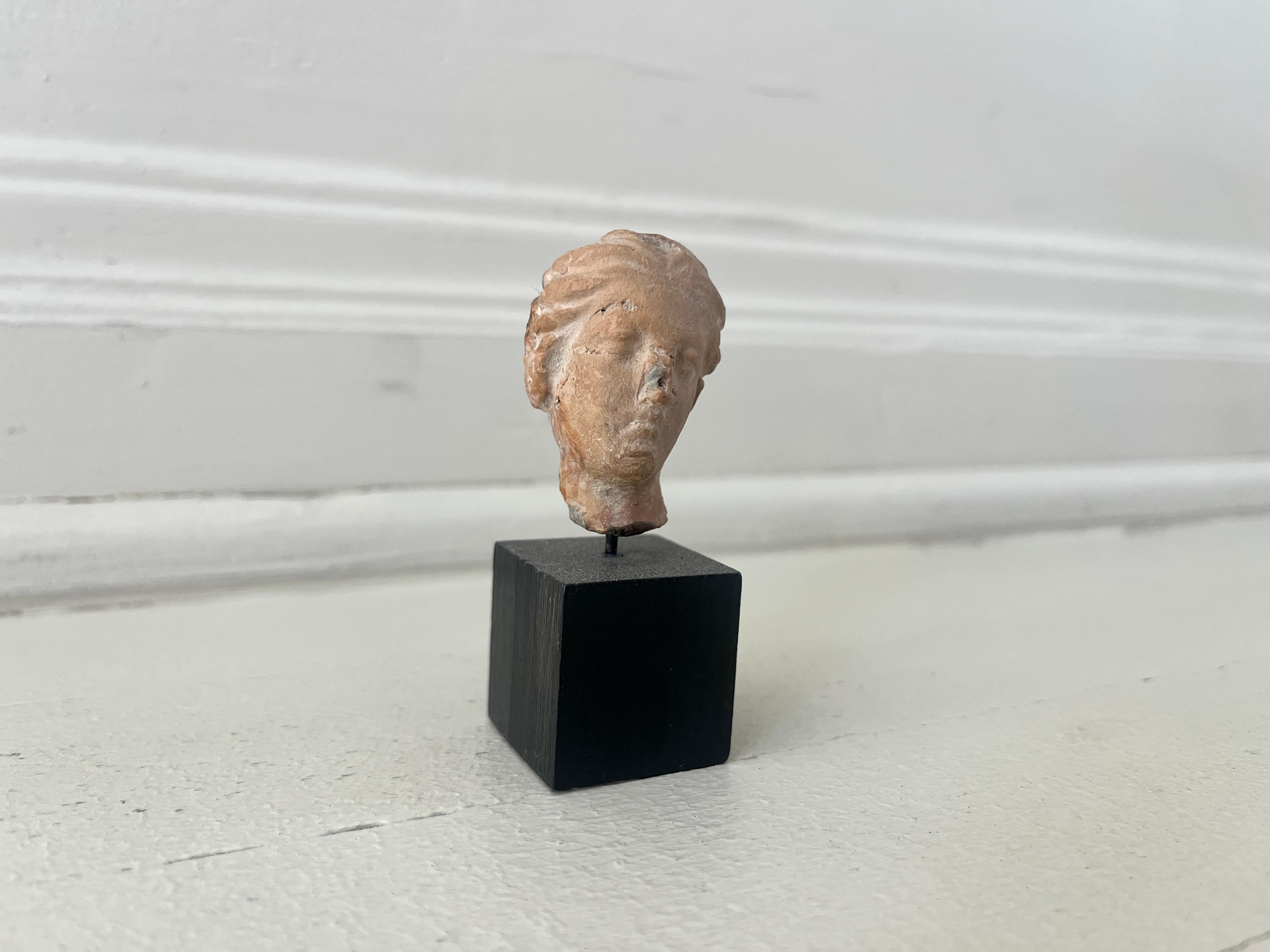 A Greek terracotta female head, c. 3rd - 2nd Century BC, her head slightly tilted to her left, features hair styled back in waves. Mounted on black wood base. Ex Royal Athena Galleries, New York. Museum Quality.