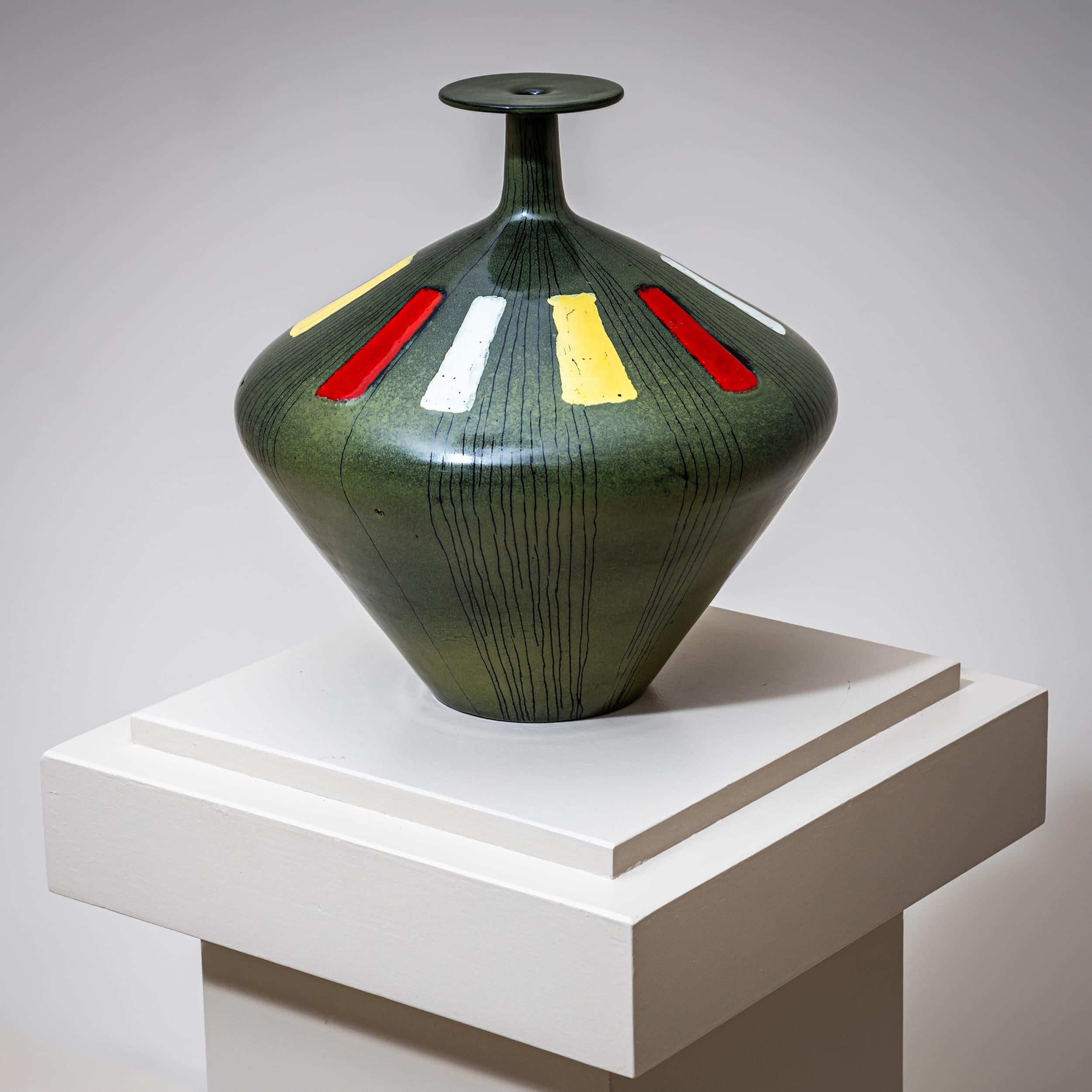 Jarrón de cerámica con esmalte verde, decoración de rayas negras y detalles rectangulares verticales en rojo, blanco y amarillo. Su característica forma bulbosa culmina en un cuello esbelto con un amplio borde en forma de disco. Marcado en la parte