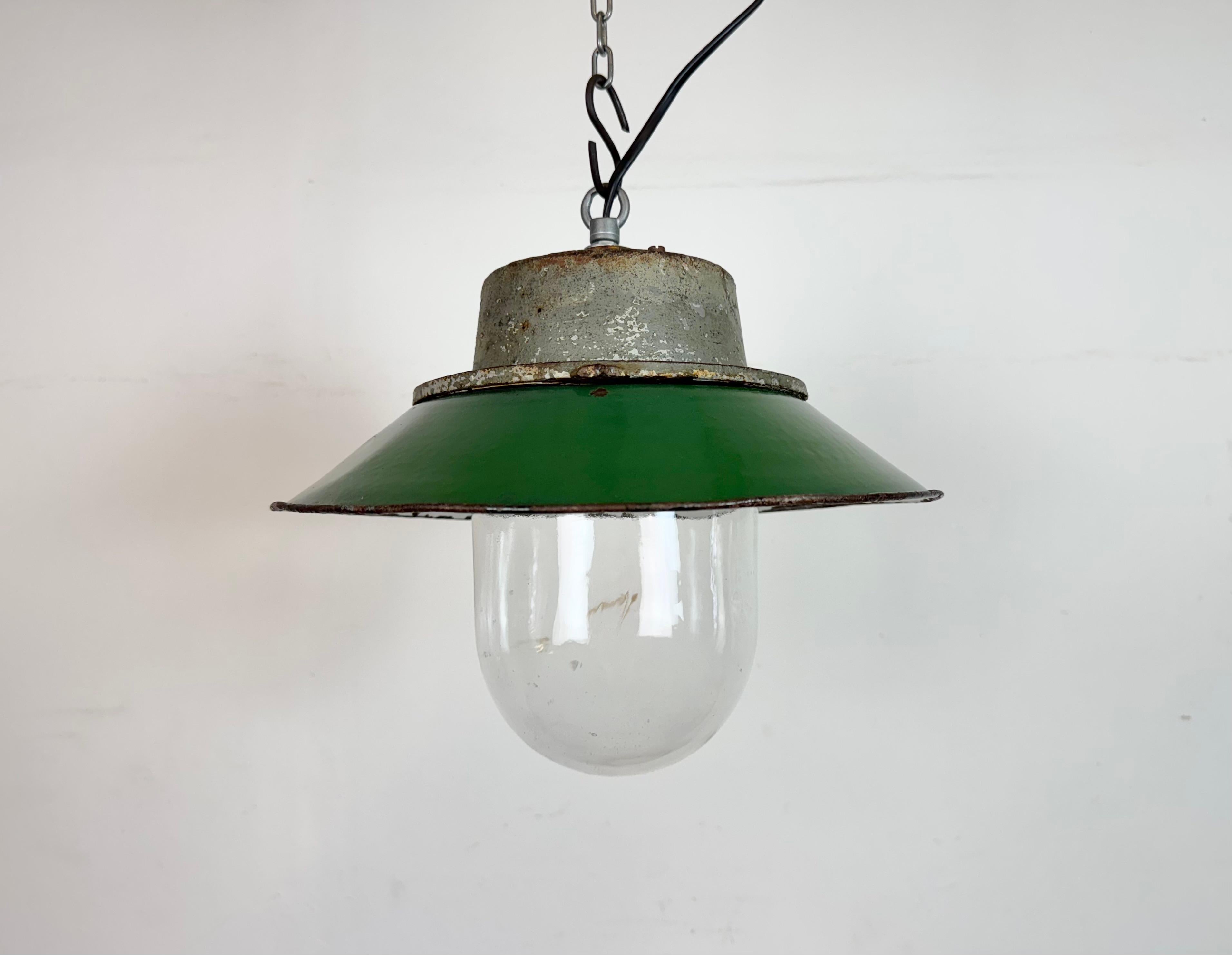 Lámpara industrial colgante fabricada en Polonia en la década de 1960. Presenta una pantalla esmaltada en verde con el interior esmaltado en blanco, una tapa de hierro fundido gris y una cubierta de cristal transparente. El casquillo de porcelana