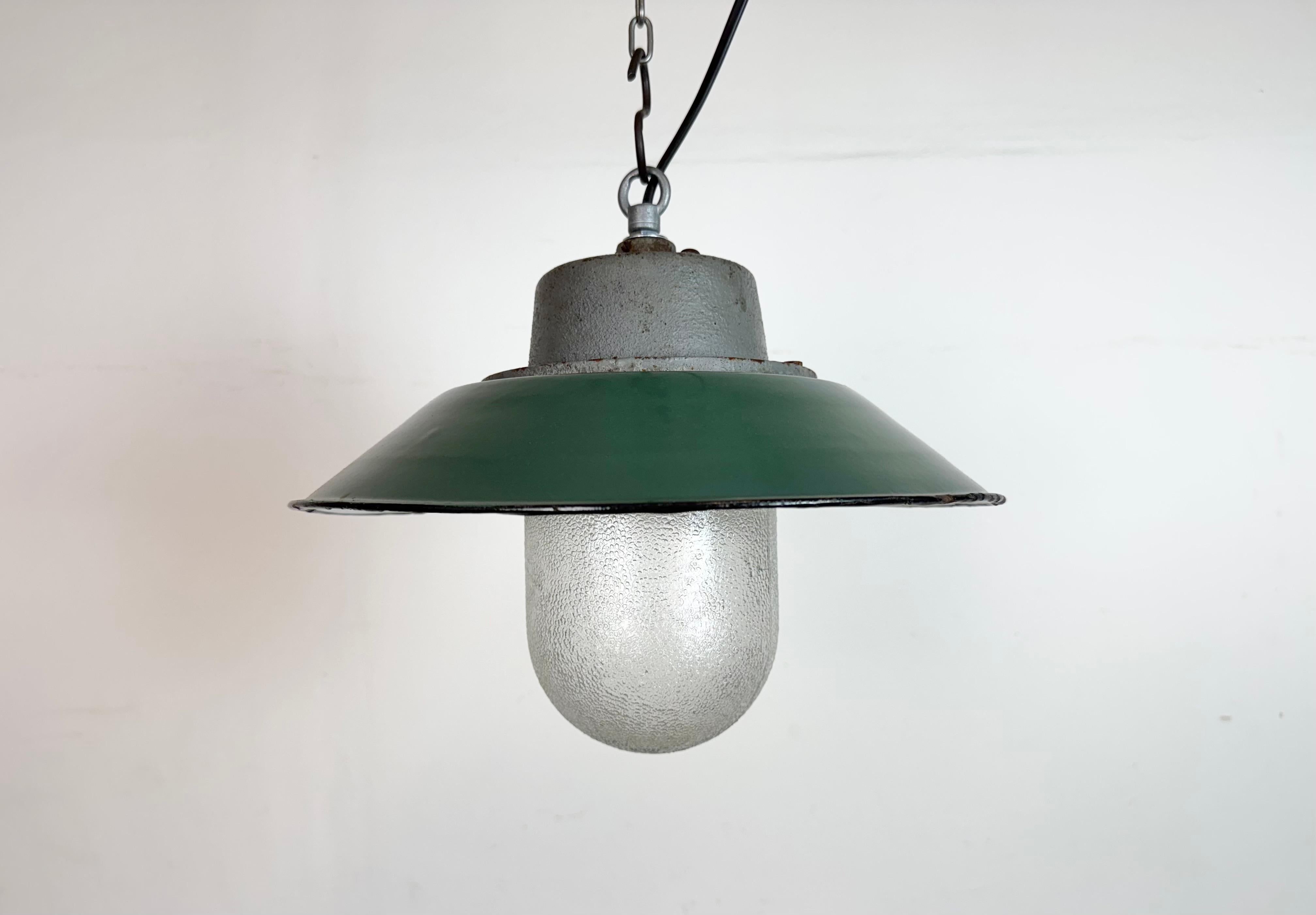 Lámpara industrial colgante fabricada en Polonia en la década de 1960. Presenta una pantalla esmaltada en verde con el interior esmaltado en blanco, una tapa de hierro fundido gris y una cubierta de cristal transparente. El casquillo de porcelana