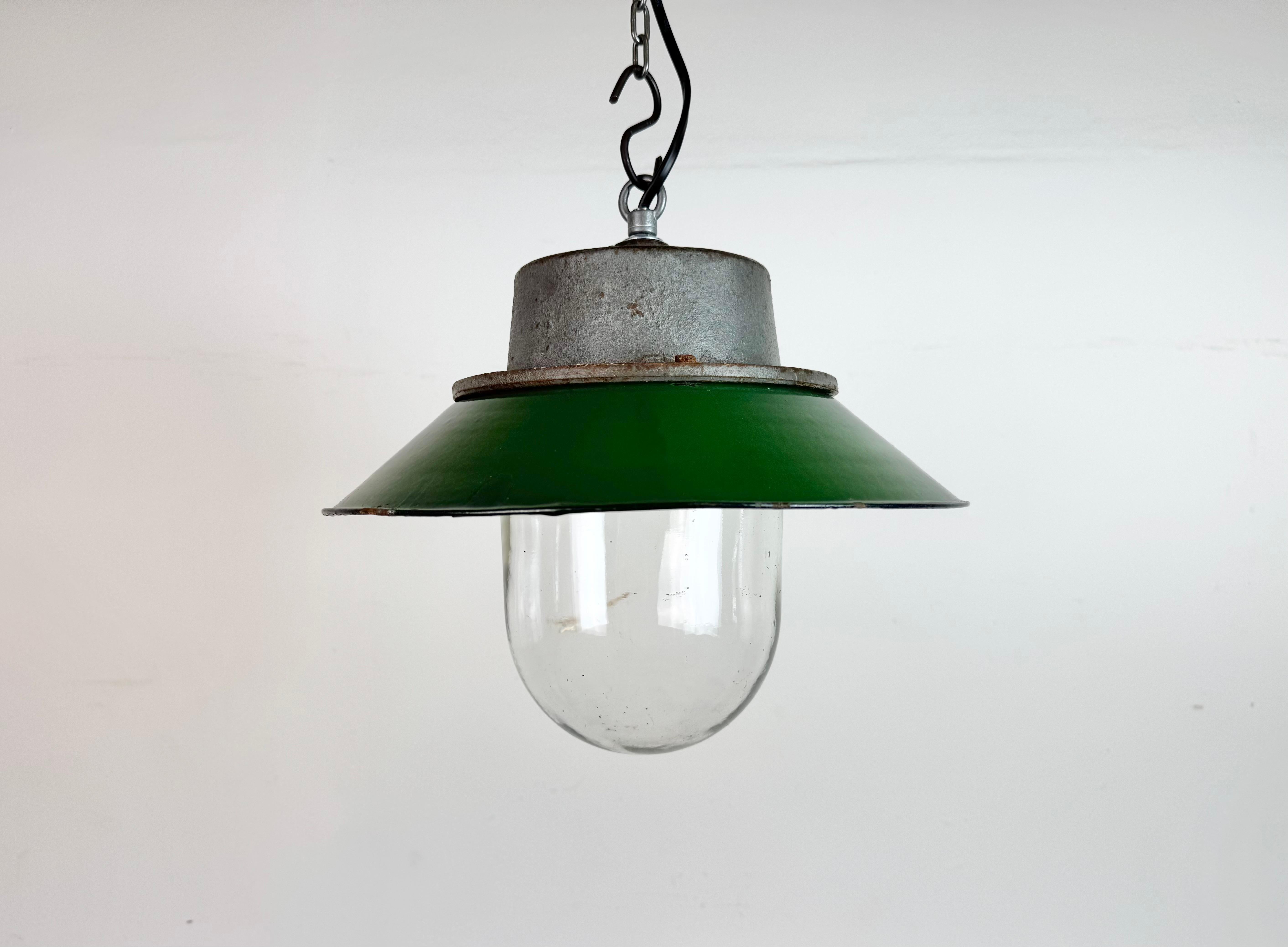 Lámpara industrial colgante fabricada en Polonia en la década de 1960. Presenta una pantalla esmaltada en verde con el interior esmaltado en blanco, una tapa de hierro fundido gris y una cubierta de cristal transparente. El casquillo de porcelana
