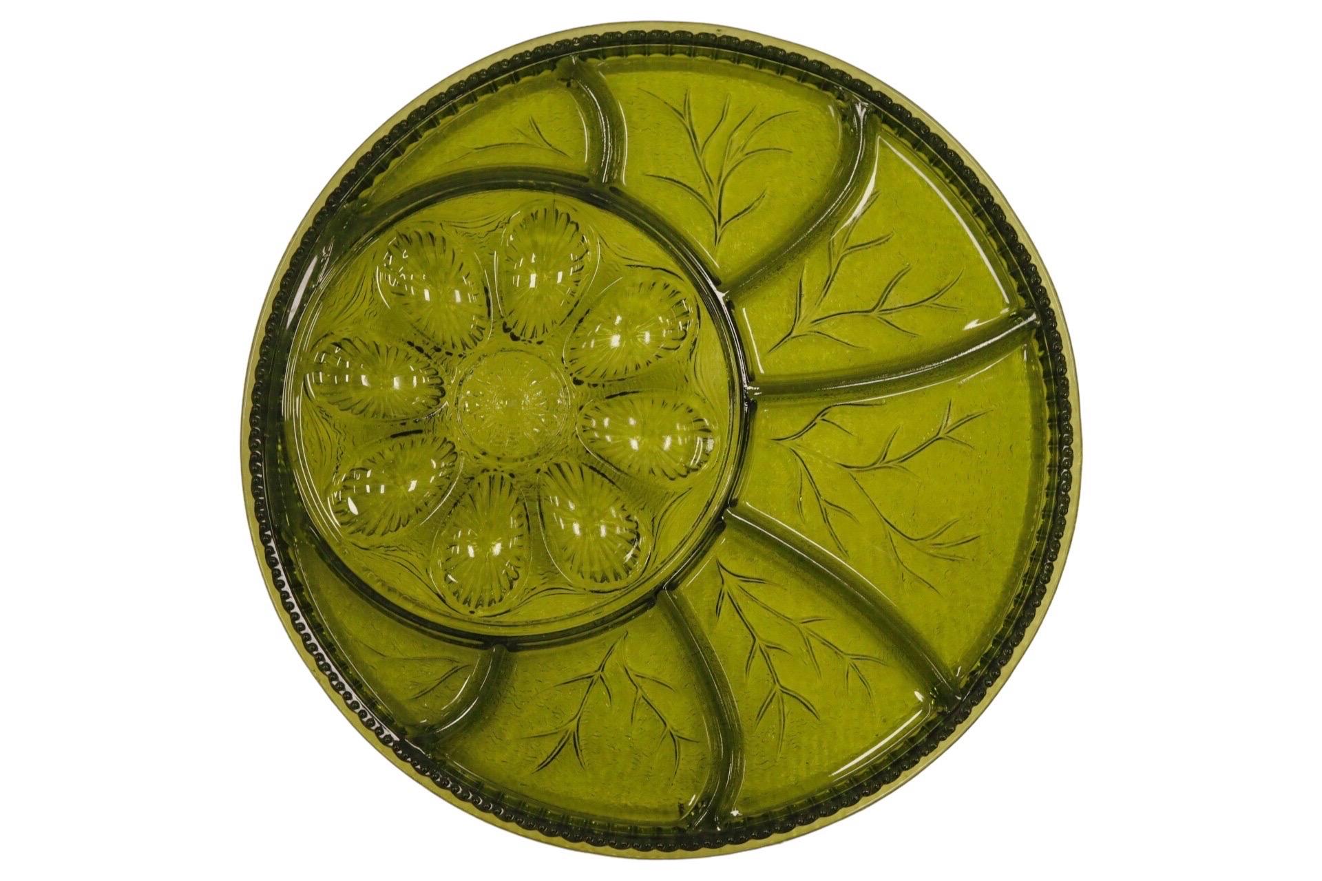 A circular glass deviled egg serving dish in avocado green. Holds eight egg halves pressed with a scalloped pattern, offset with seven additional serving compartments pressed to give a veined leaf look. Finished with a ribbed edge.
