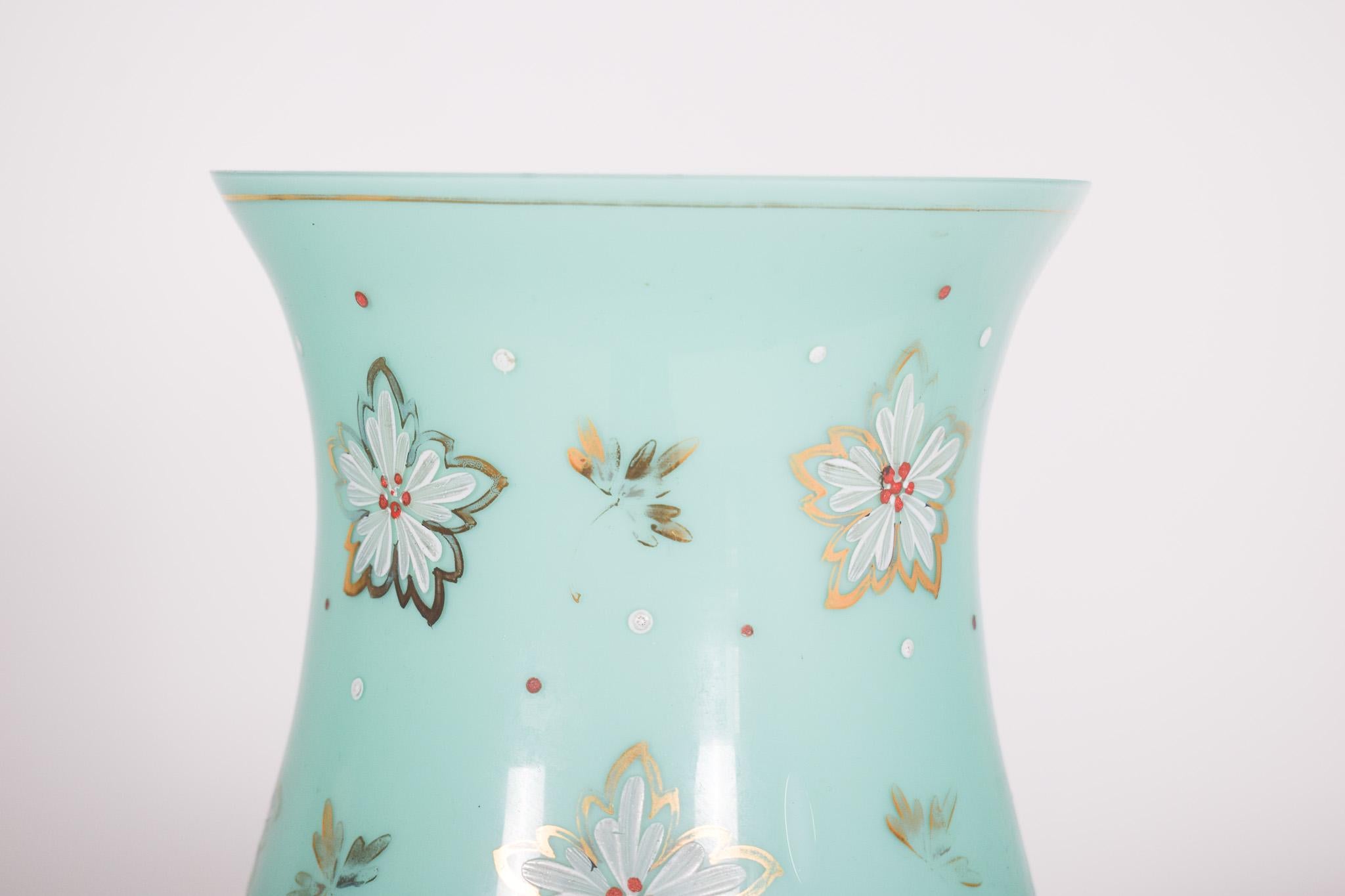 Vase en verre vert avec des fleurs. Décor polychrome et or, avec fleurs, feuilles et éclaboussures. Une frise d'or sur la bouche et deux sur la base. De forme incurvée à l'ourlet, elle s'affine légèrement avant de s'ouvrir sur le haut, offrant une
