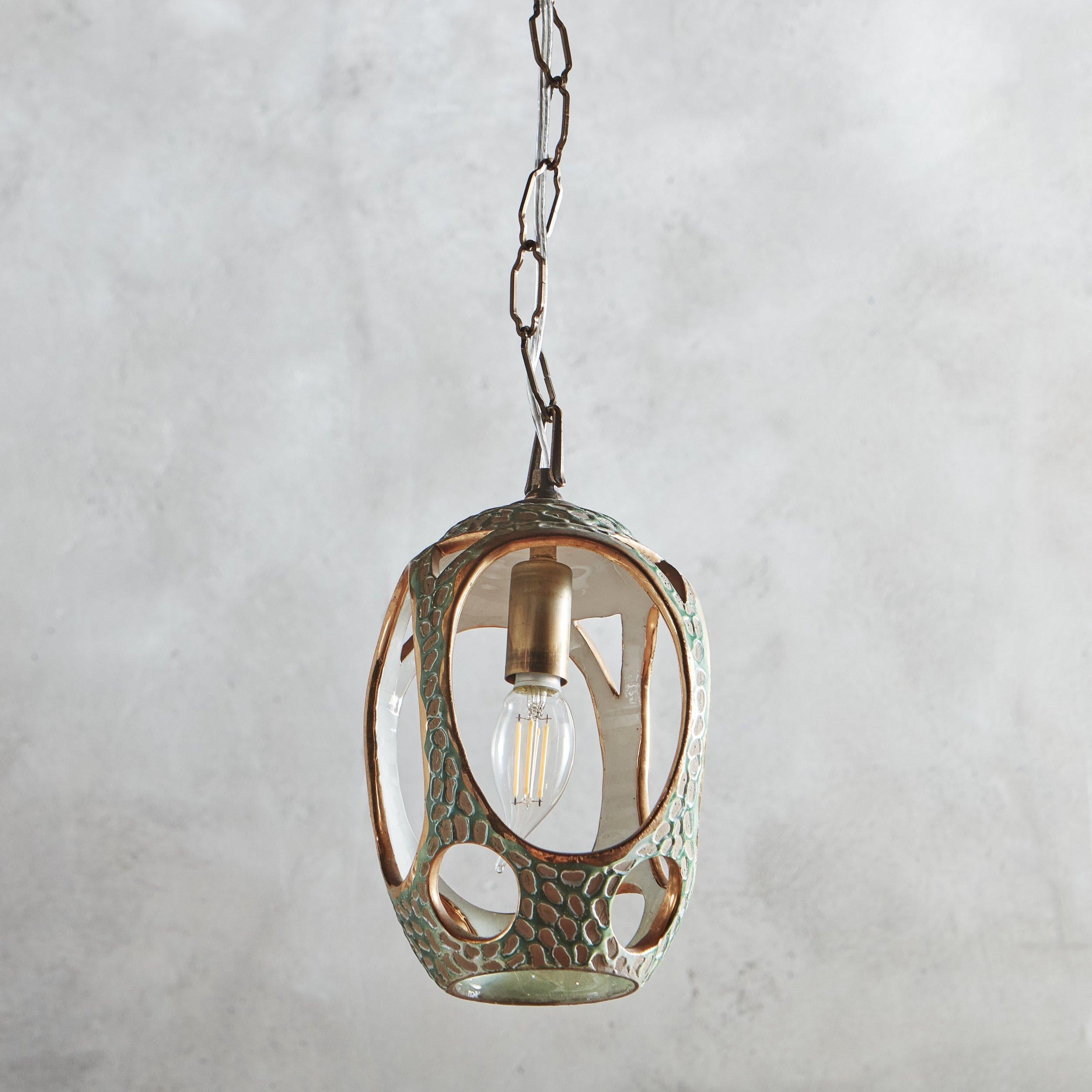 A 1950s glazed ceramic pendant light with oval and circular cutout details on the sides and base. This fixture has gold detailing and a textural exterior in a vivid green hue, which contrasts beautifully with the reflective white interior. This
