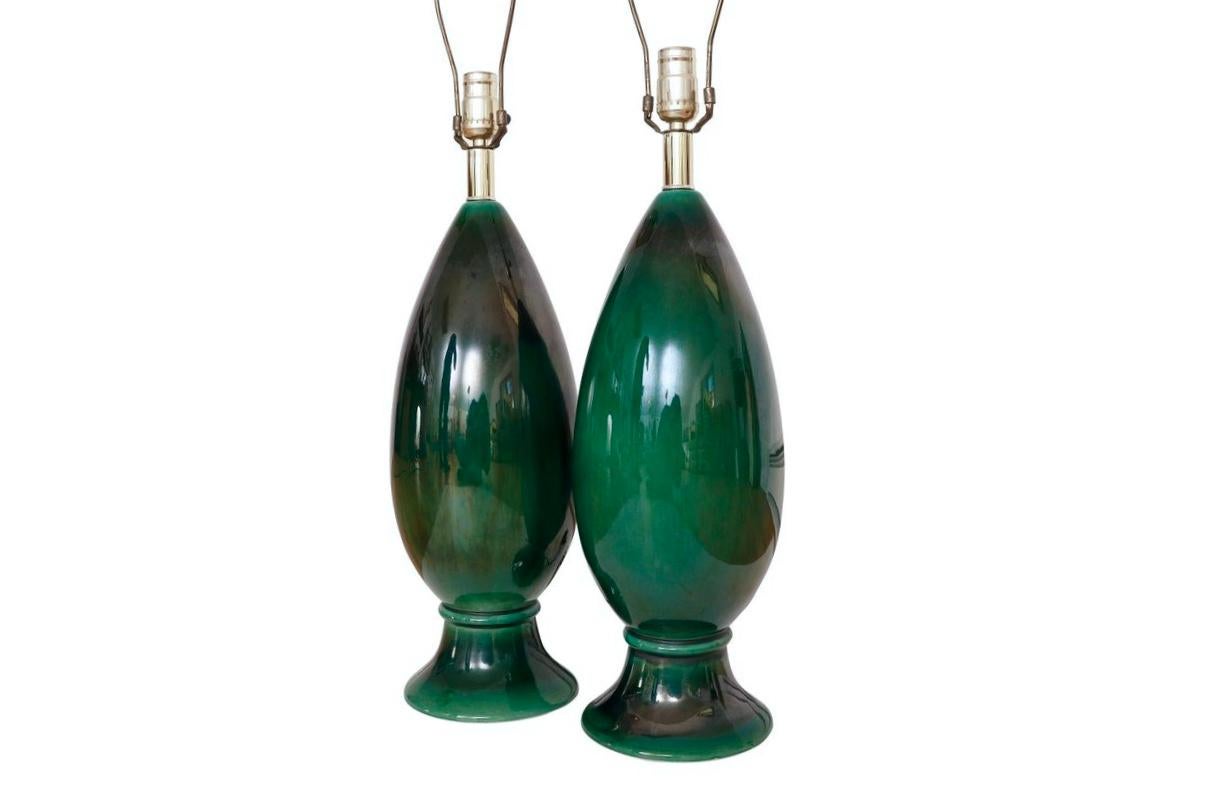 A pair of mid century oval ceramic table lamps glazed a bright bottle green. Decorated with a beveled ring above a round foot. A short brass column supports the bulb and harp. Each lamp measures 6