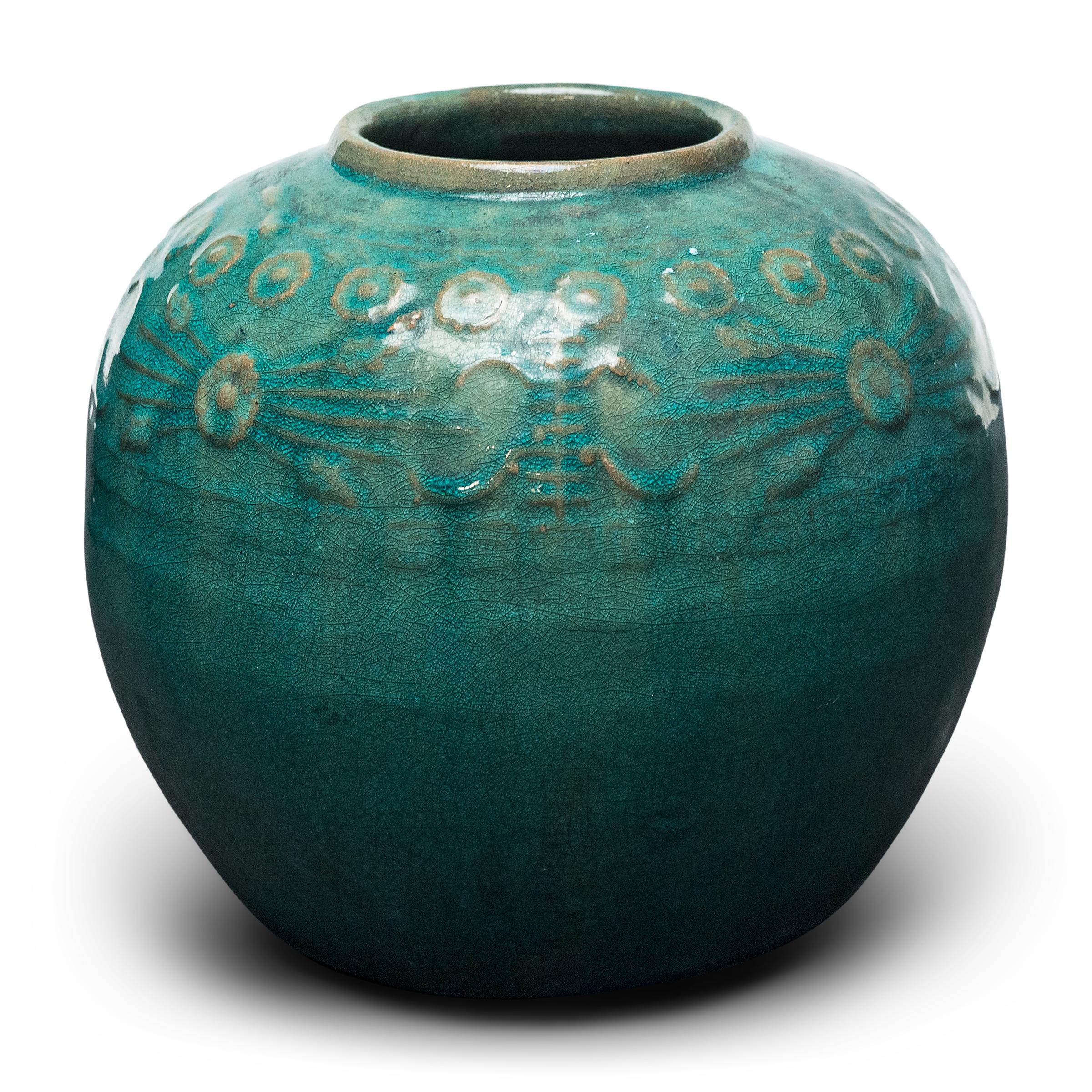 A glassy, blue-green glaze sheets, pools, and drips across the rounded body of this provincial kitchen pot, lingering on the low relief decoration to the jar's shoulders. Among the raised patterns are round chrysanthemum blossoms and stylized Shou