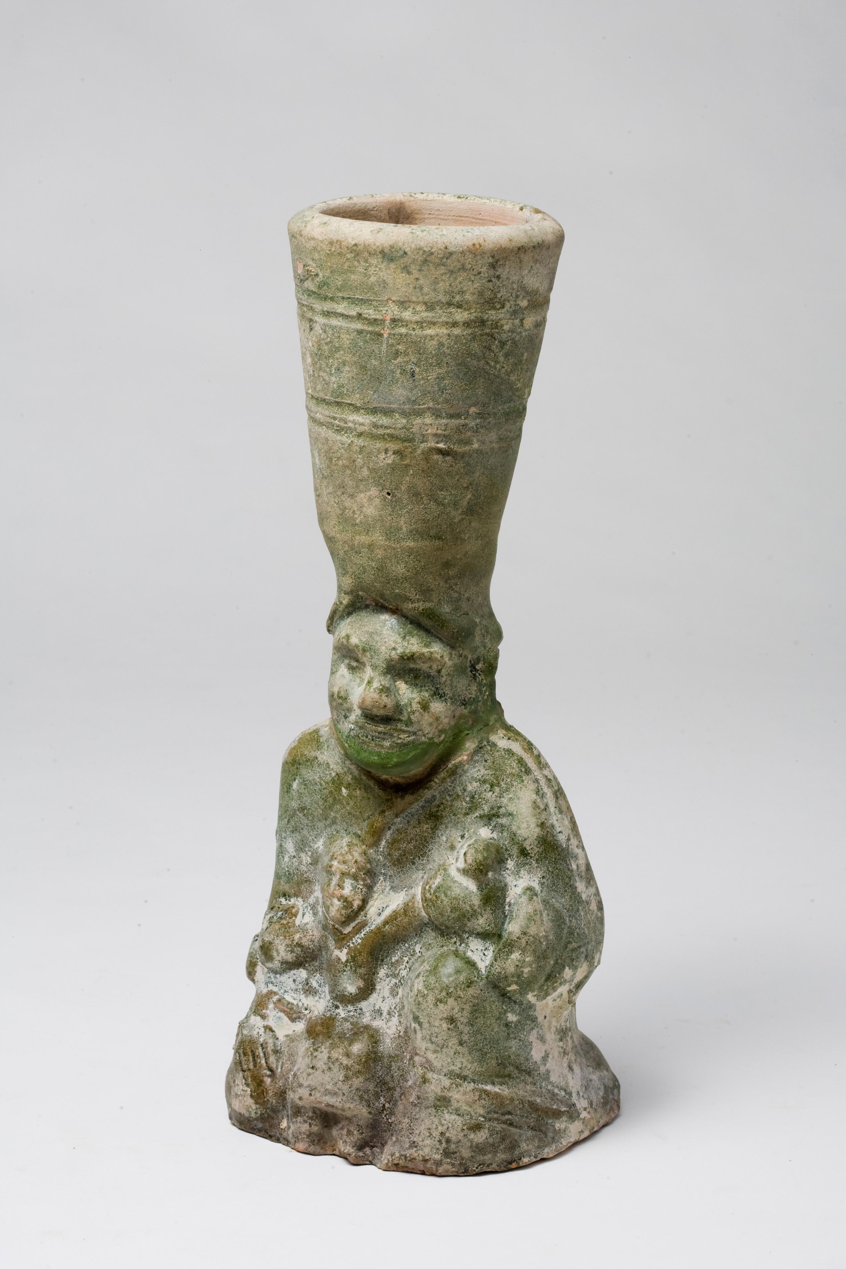 The lamp shaped as a kneeling female figure wearing long robes, her face with a large nose, deep-set eyes and pronounced cheeks, holding an infant in her lap, her cylindrical and elongated headgear forming the cupped oil holder, wood stand.

Period