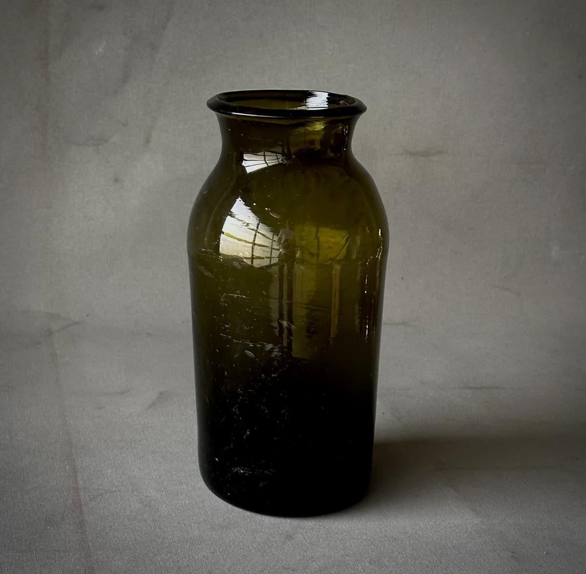 Questa antica bottiglia francese in vetro soffiato a mano, risalente alla metà del XIX secolo, affascina per la sua ricca tonalità olivastra e il suo fascino rustico. Realizzato artigianalmente, ogni pezzo di questa collezione di undici pezzi vanta