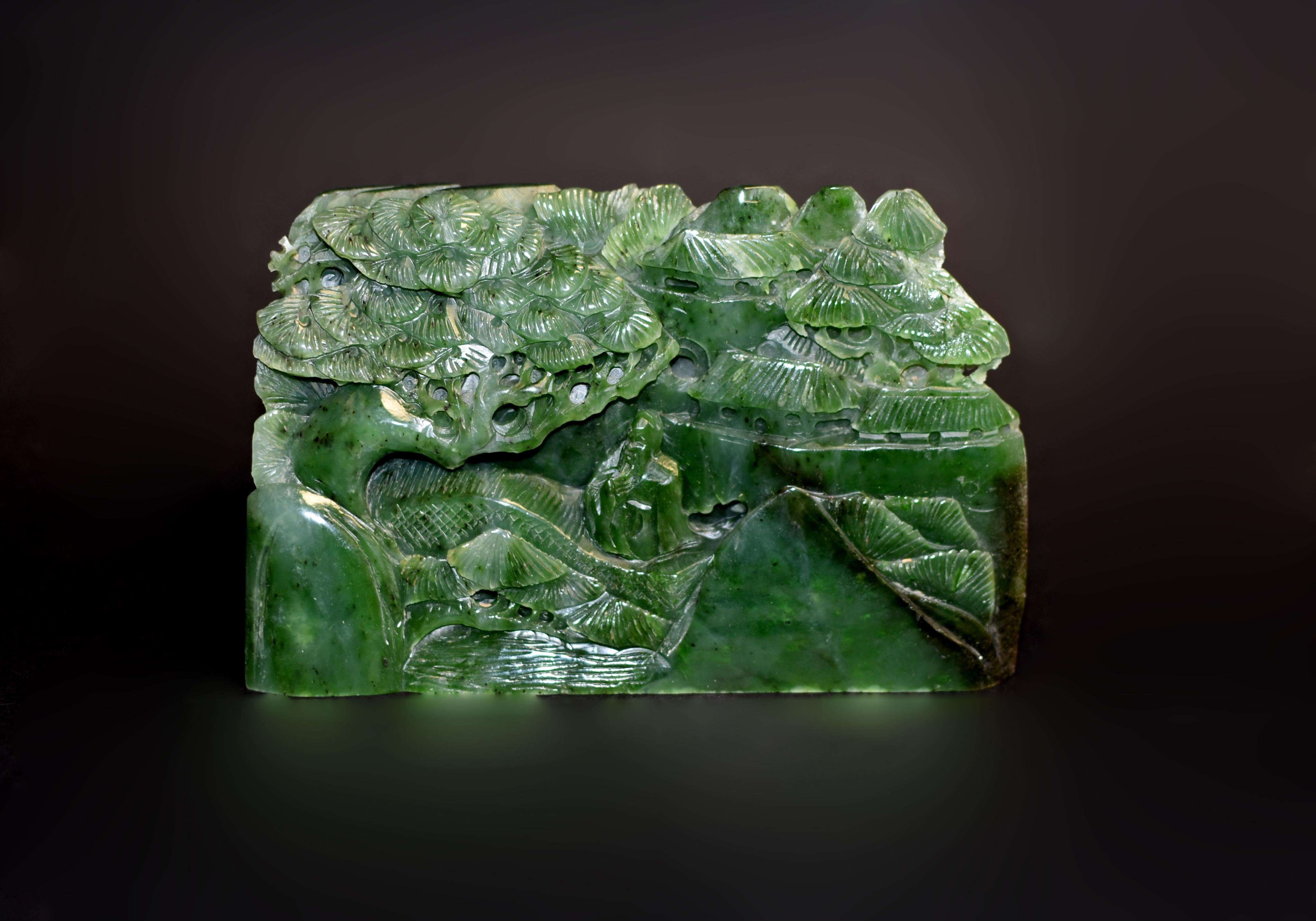 A fine sculpture of solid nephrite jade depicting a peaceful lifestyle. The three dimensional piece features rolling hills and a prominent Chinese pine tree in the foreground, an elderly scholar crossing a bridge with smooth running water underneath