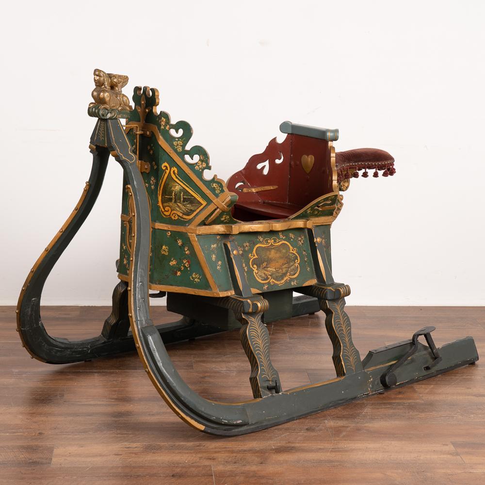 Highly decorative and elaborate green painted single horse drawn sleigh.
Pictorial gold trimmed panels, flowers, and a gold painted griffin crowning the front are just of few of the elements that make this sled such a special find. 
A red velvet