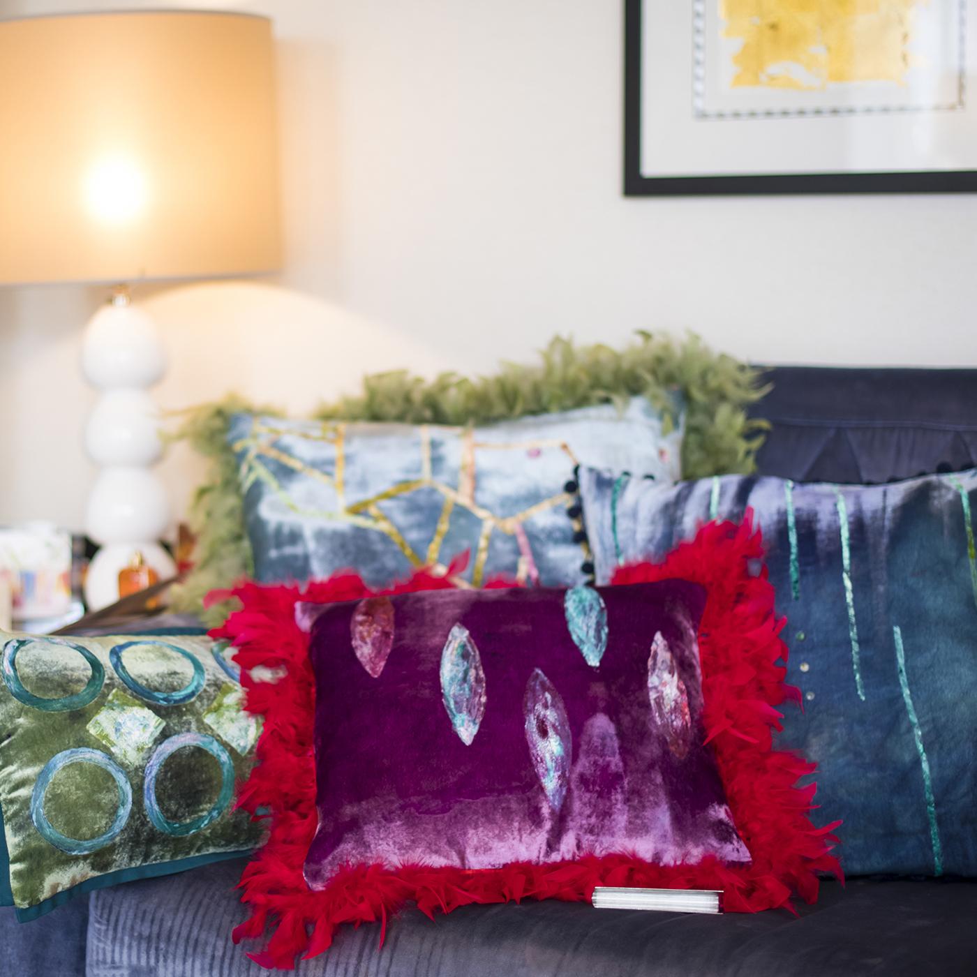 A geometric motif dominates the front of this sophisticated silk velvet pillow, captured in multicolored nuances on a shimmering blue background whose soft texture is recalled in the delicate fringe of green feathers. The rich and saturated colors