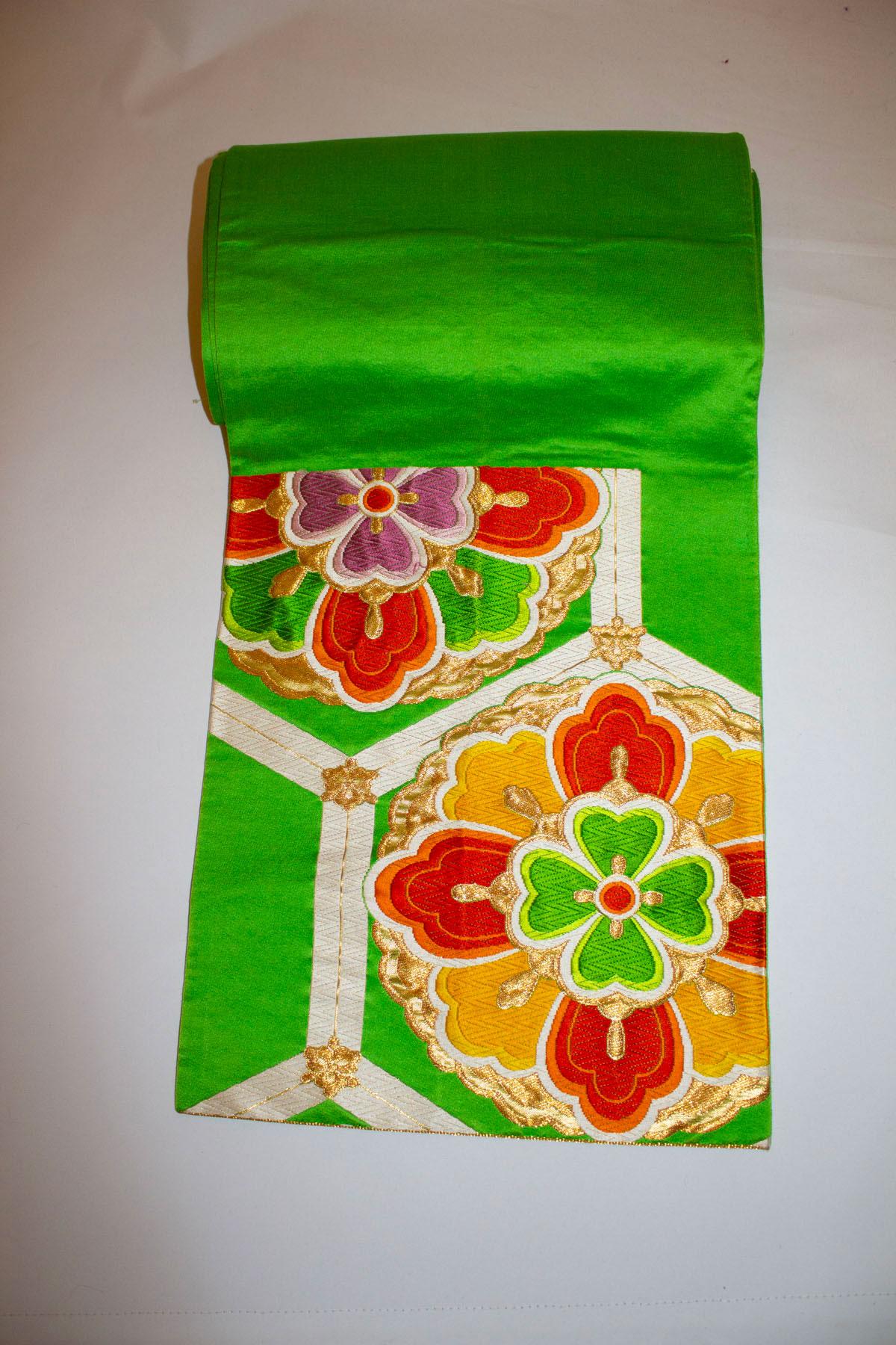 Ein wunderschöner Obi-Gürtel für den Frühling. Der Gürtel hat einen leuchtend grünen Hintergrund und ein sechseckiges Muster mit grünen, violetten und orangefarbenen Blumen. Gesamtlänge 12'' x 162'', gestickte Bahnen messen. 12'' x 14'' und 12'' x