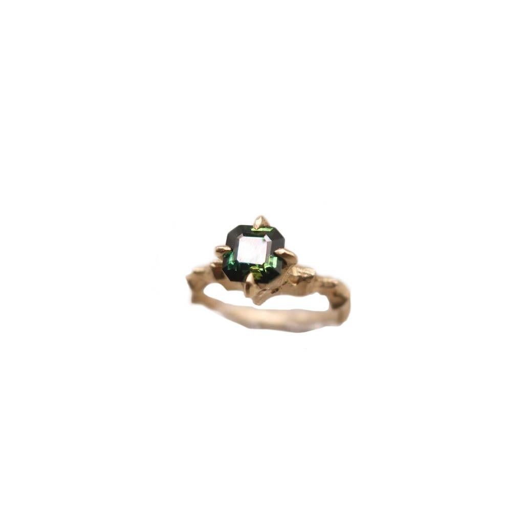 A beautiful asscher cut green sapphire is hand set in 14 Karat yellow gold. This solitaire style features a pointed prong claw- like setting and detailed band. This piece is hand carved from wax and the shank displays ridges and peaks in an
