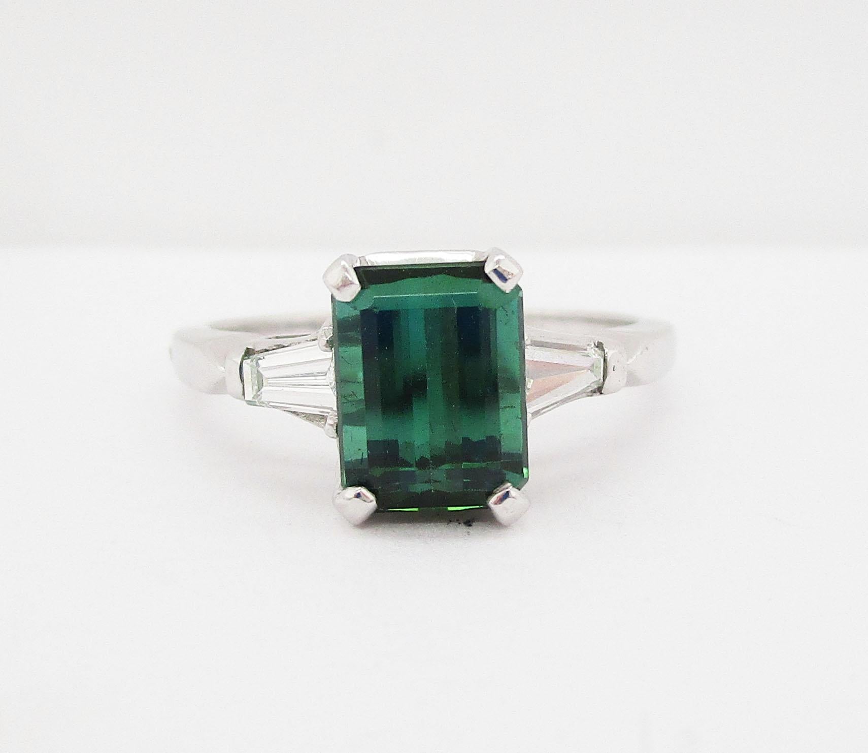 This is a gorgeous white platinum ring featuring an emerald cut green tourmaline center flanked by brilliant white tapered baguette diamonds. The center stone has a rich, deep green hue and is incredibly well cut, creating a dimensional, sparkling