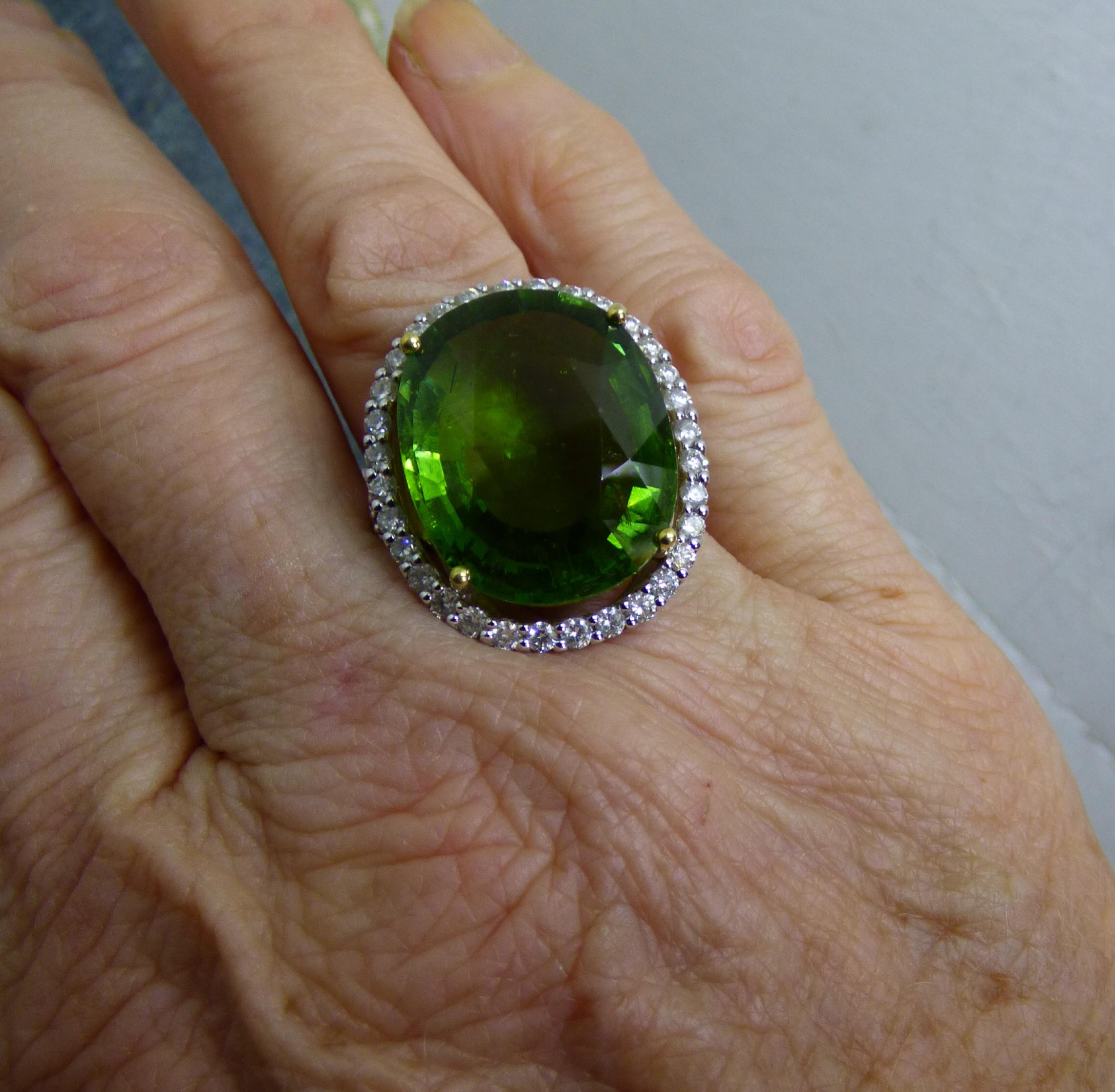 Clean and bright green Tourmaline oval cut stone (22.12ct) is surrounded by Diamonds with Diamonds set on the side shanks. Total Diamond weight is 1.70ct.   The total size of the cluster is 20X16mm. The ring is handmade in 18K white gold with yellow