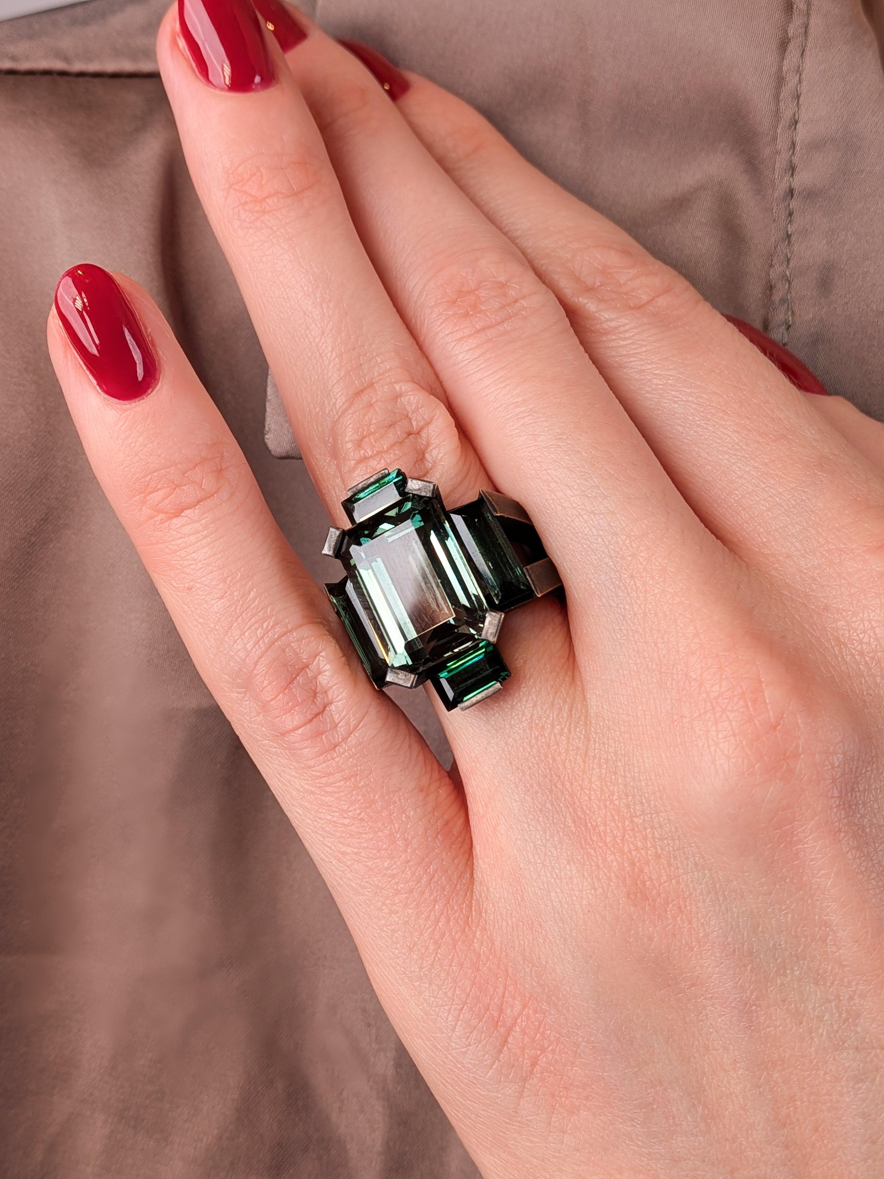 Anillo de turmalina verde en bronce ennegrecido con una piedra central de aproximadamente 8 quilates y turmalinas adicionales engastadas en un diseño geométrico en cruz, de líneas arquitectónicas marcadas y estética escultural contemporánea.

Este