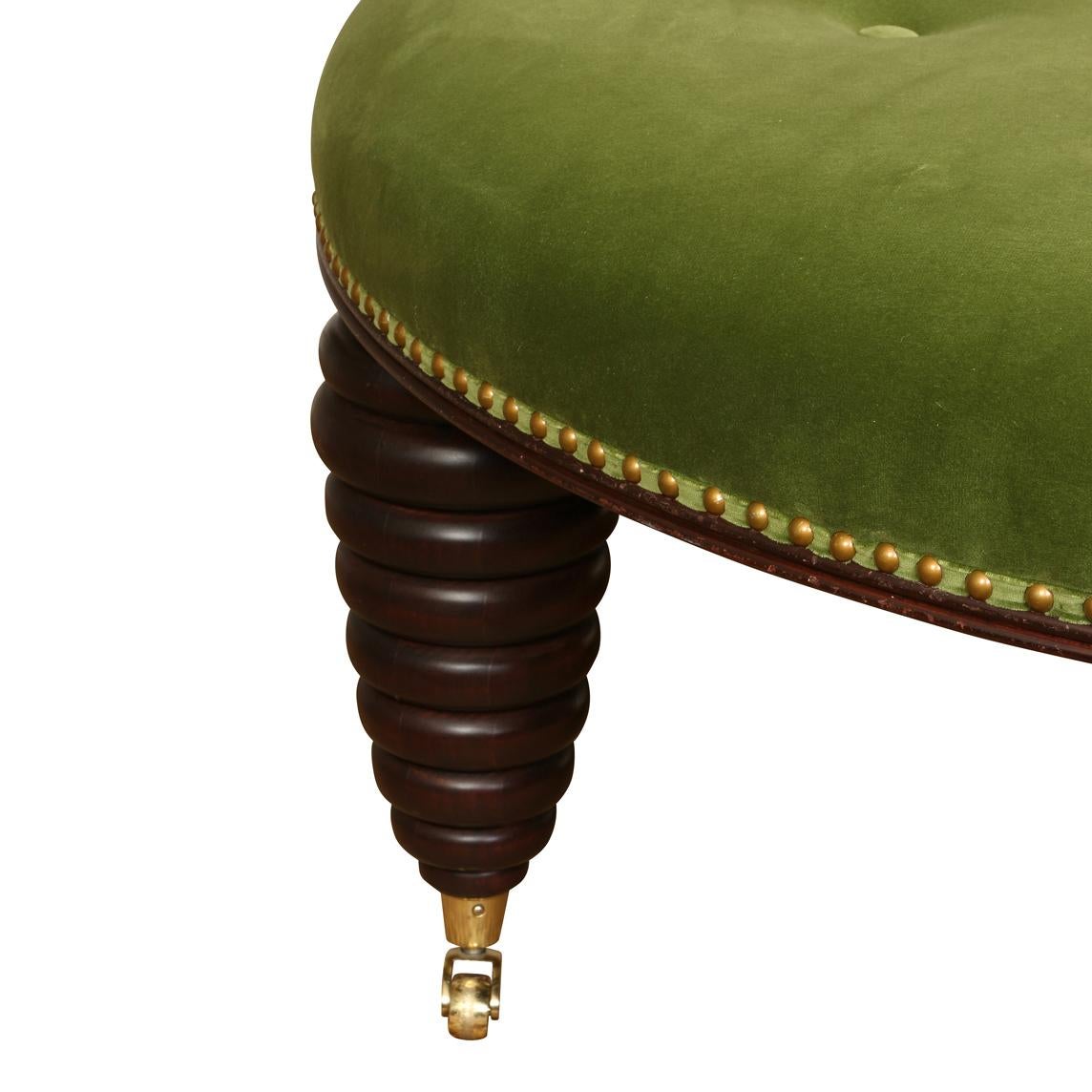 A generously sized, round button tufted ottoman with nailhead trim, newly reupholstered in green velvet with stacked, circular modern shaped legs on casters