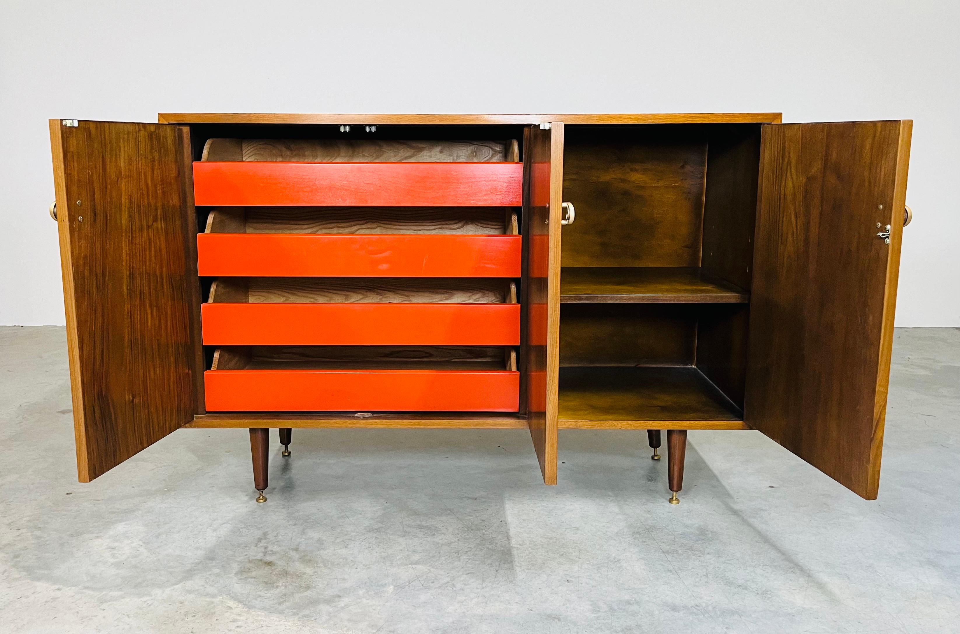An elegant Mid-Century Modern credenza having walnut construction with 4 orange internal drawers to the left and a case with adjustable shelf to the right having curved brass pulls designed by Greta Magnusson-Grossman for Glenn of California Circa