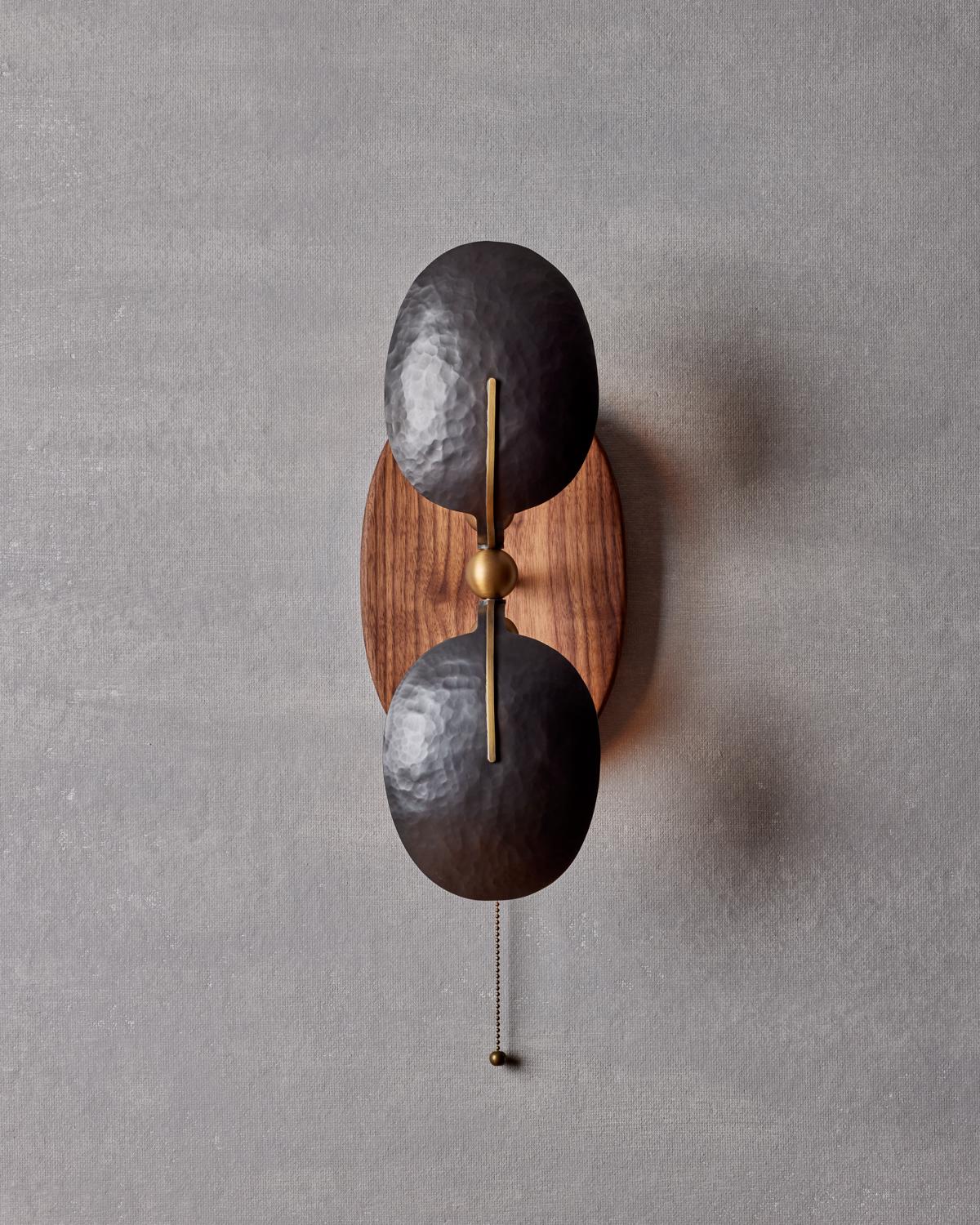 The Greta Sconce mounted from a slightly concave walnut backplate is accented with two bench-made bronze reflectors and oval brass sockets.

Materials:
Wood, Bronze, Brass

Fixture Finishes: Walnut, Light Antique Brass, Hand Hammered Bronze