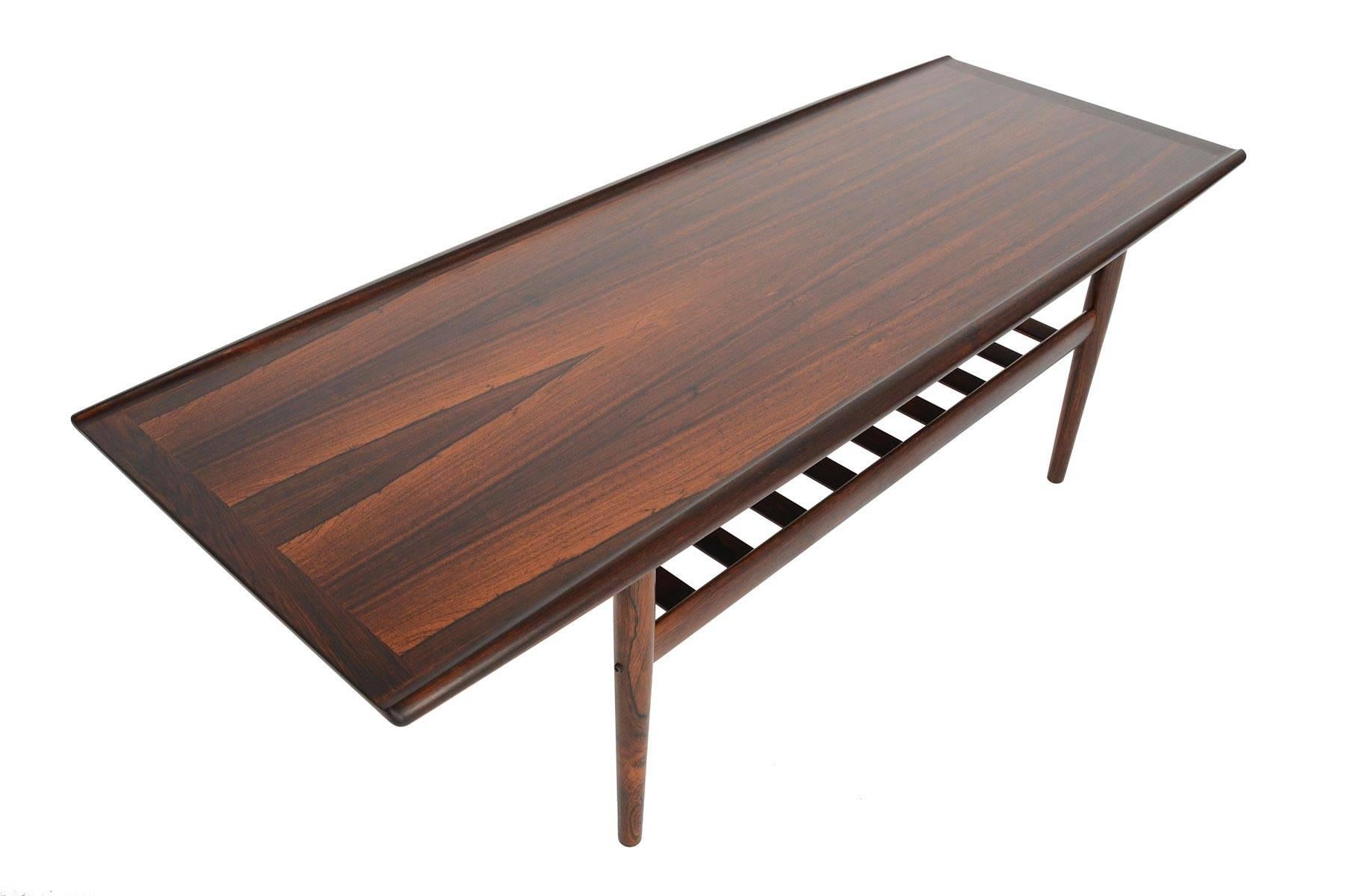This elegant Danish modern surfboard coffee table was designed by Grete Jalk for Glostrup Mobelfabrik in the 1960s. Beautiful book-match veneer is flanked by raised lips. A lower rack offers storage. Recently refinished and in excellent condition.