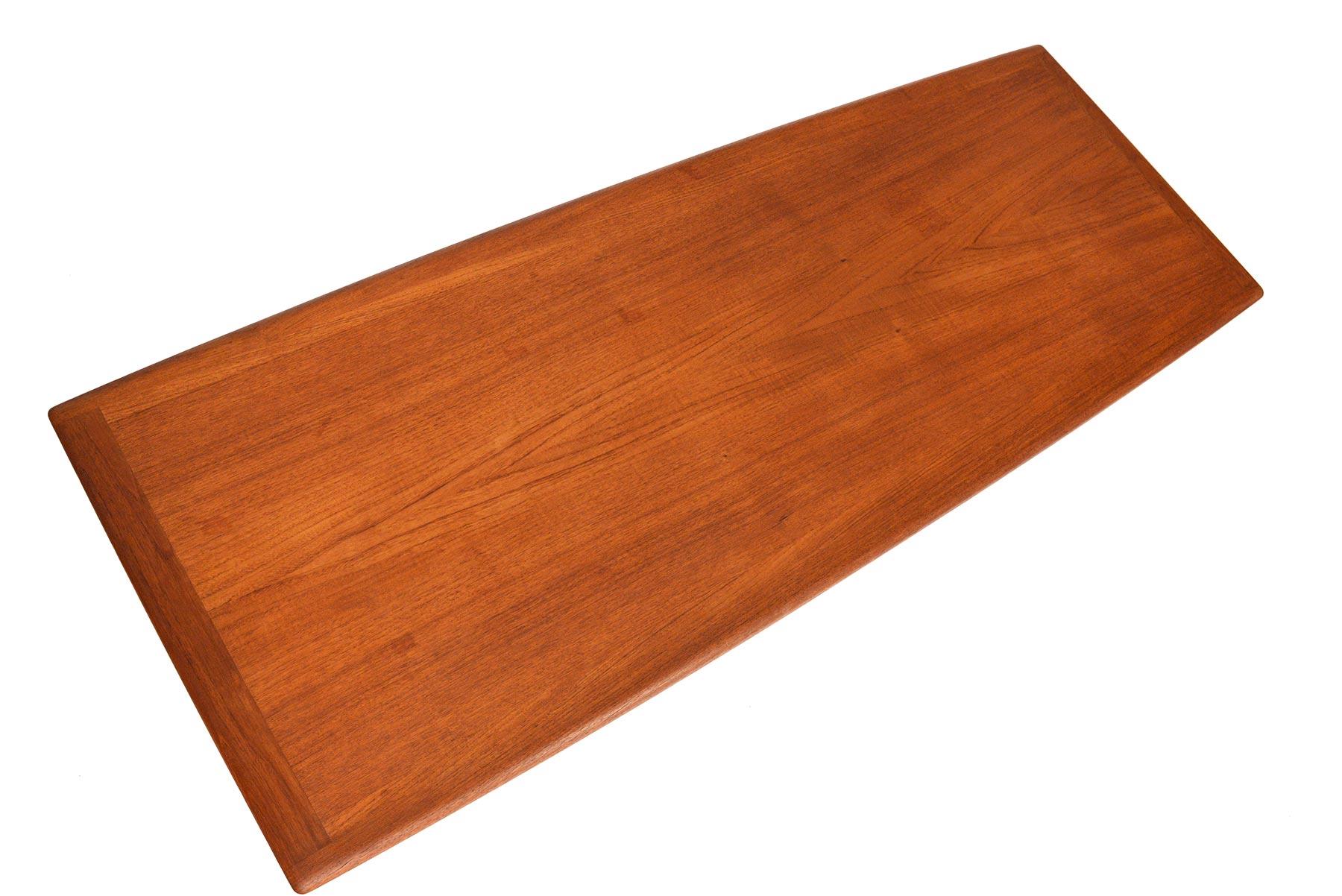 A staple of Scandinavian design, this teak surfboard coffee table was designed by Grete Jalk for Glostrup Møbelfabrik in the 1960s. A large teak table surface features a curved lip on the underside. A slatted rack sits below for storage. In