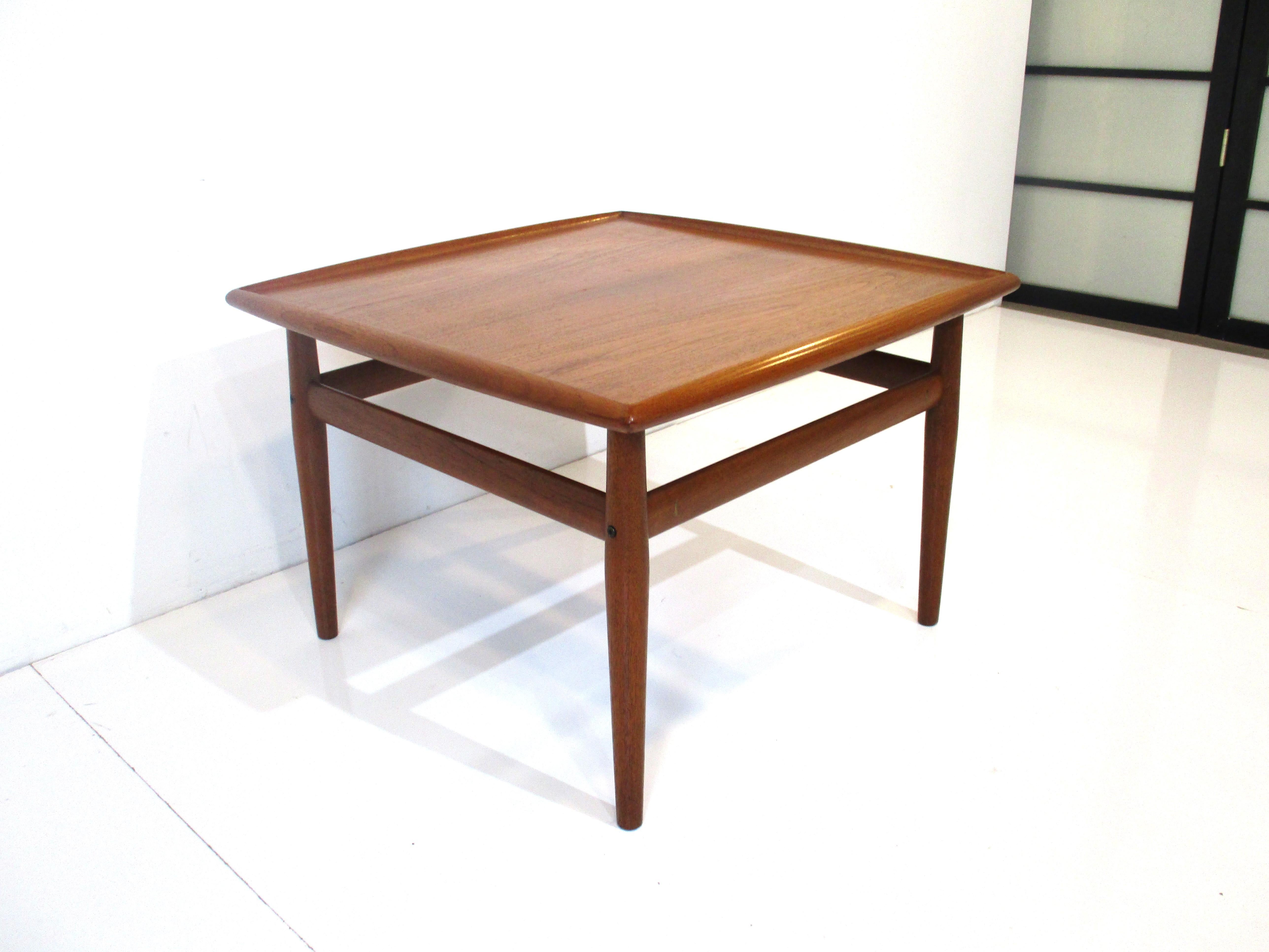 A teak wood coffee table with very well crafted upper lip to the top and stretchers to each leg having brass details . Manufactured in Denmark and designed by the iconic female designer Grete Jalk.