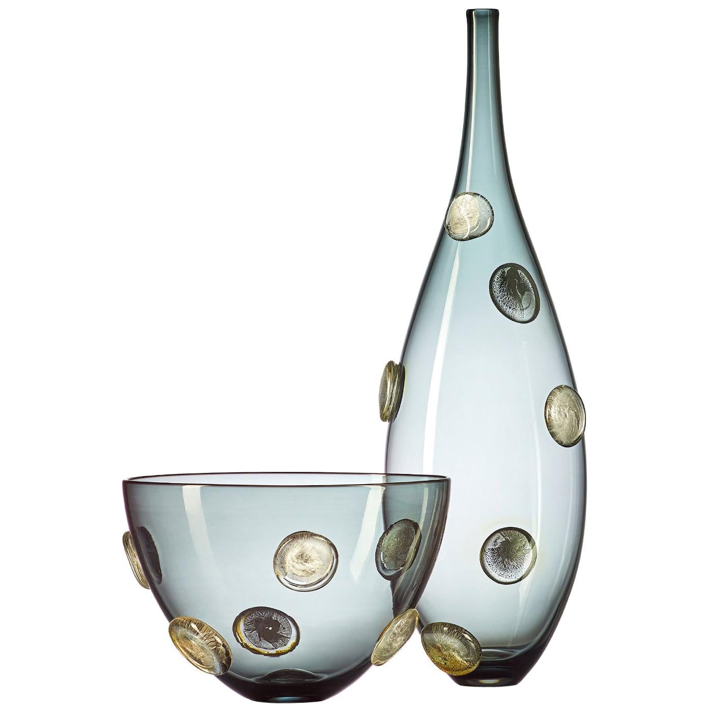A luminous statement in translucent grey hand blown glass, the Moneta bowl with silver dots features a Cascade of raised metallic coin-shaped dots, encased in clear glass. This large scale designer vase is hand blown from start to finish by Vetro