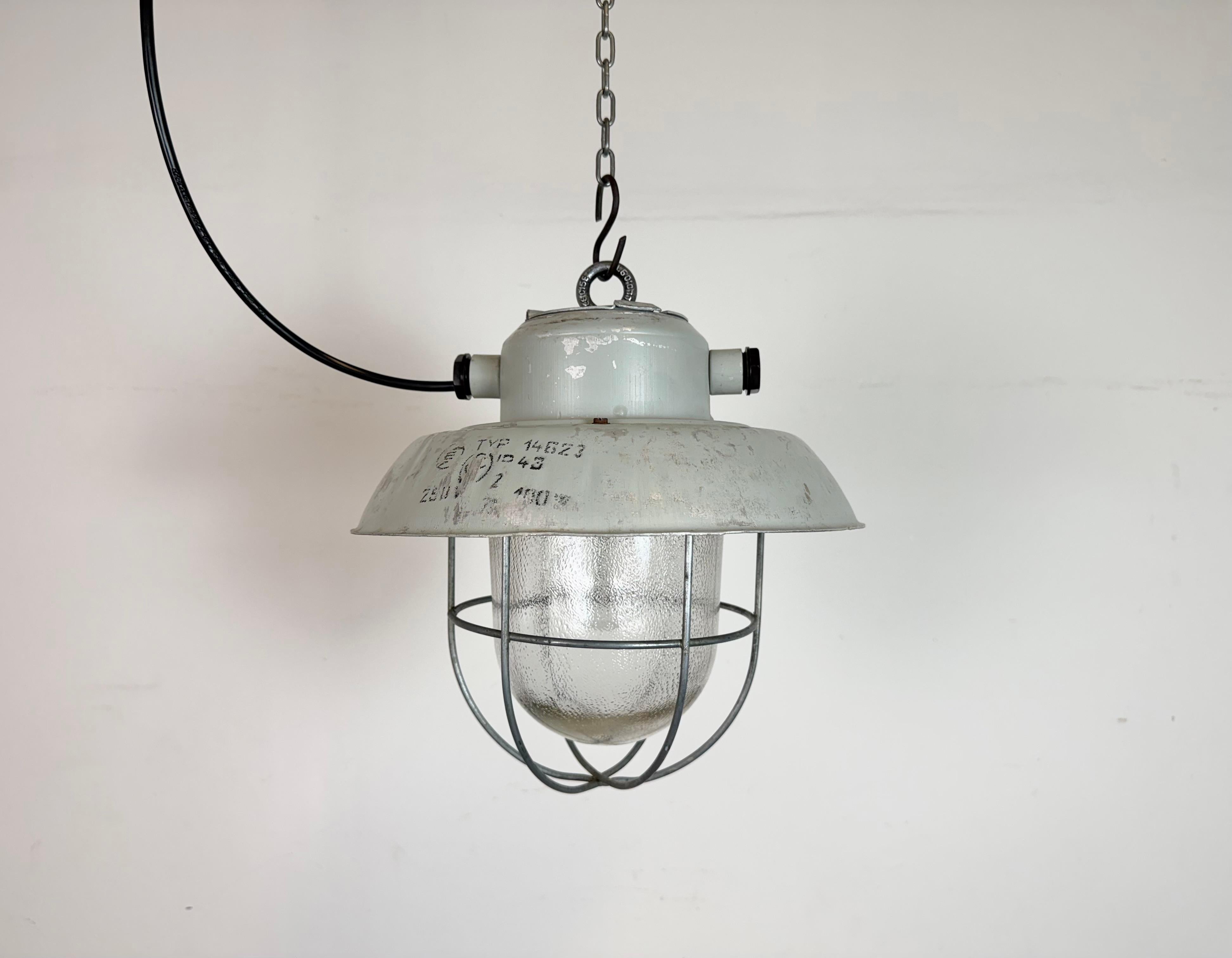 Lámpara colgante de fábrica industrial fabricada por Elektrosvit en la antigua Checoslovaquia durante la década de 1970. Presenta una pantalla de aluminio gris, un cristal esmerilado y  una rejilla de hierro. En  El casquillo de porcelana requiere