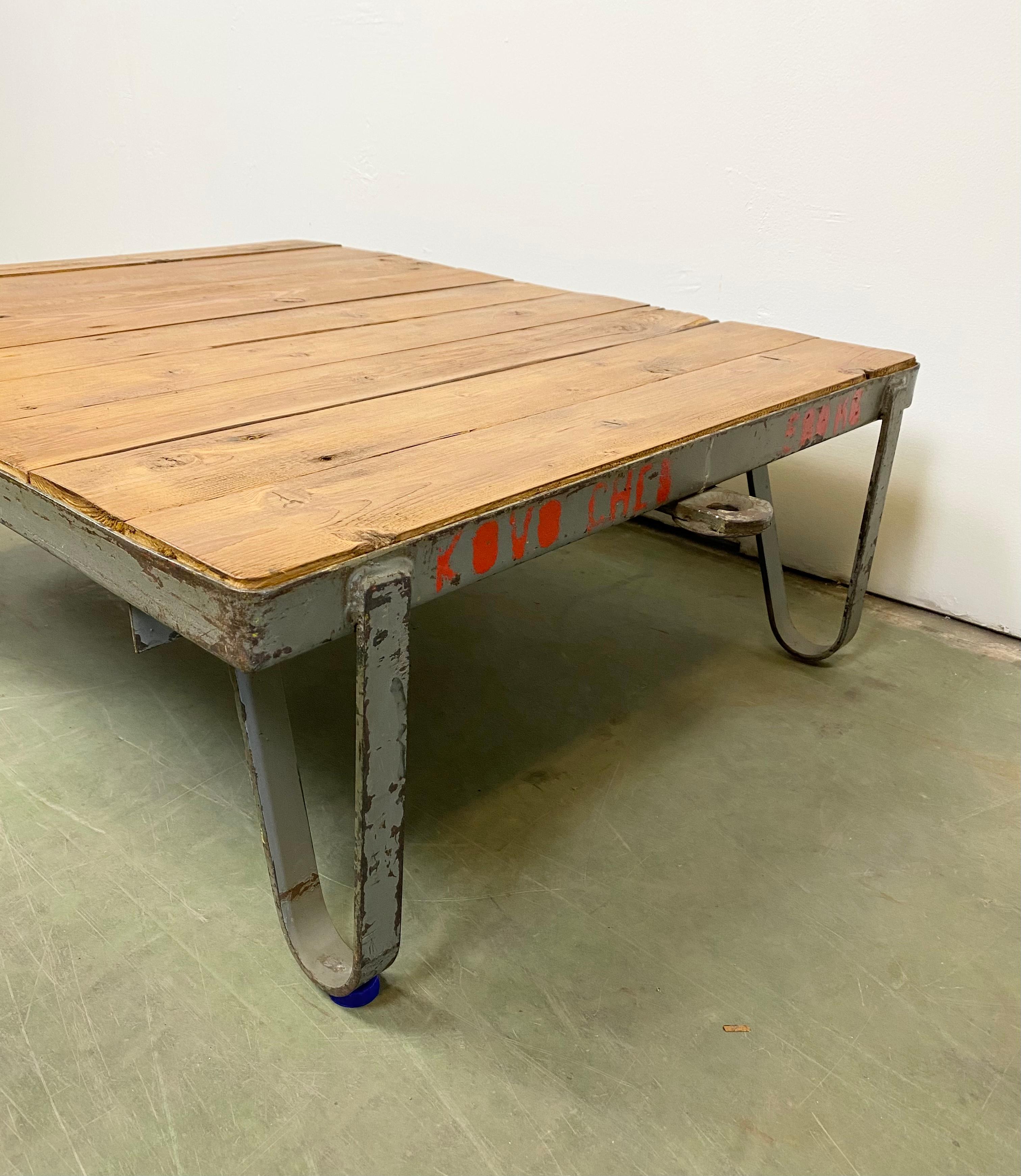 Former pallet truck from a factory now serves as a coffee table. It features a grey iron construction with two wheels and a solid wooden plate. The weight of the table is 30 kg.