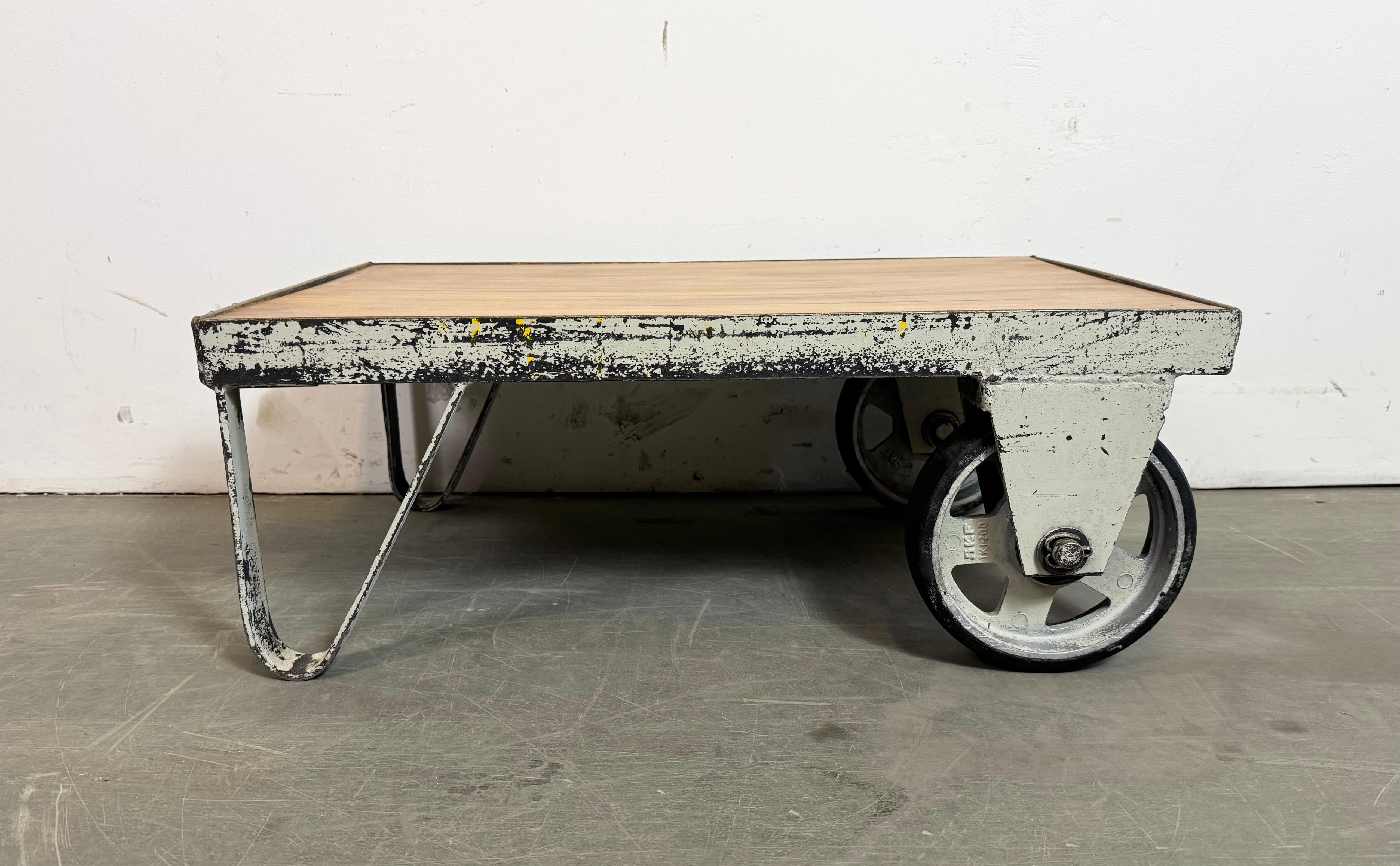 Former pallet truck from a factory now serves as a coffee table. It features a light grey iron construction with two original wheels and a solid wooden plate. The weight of the table is 26 kg.