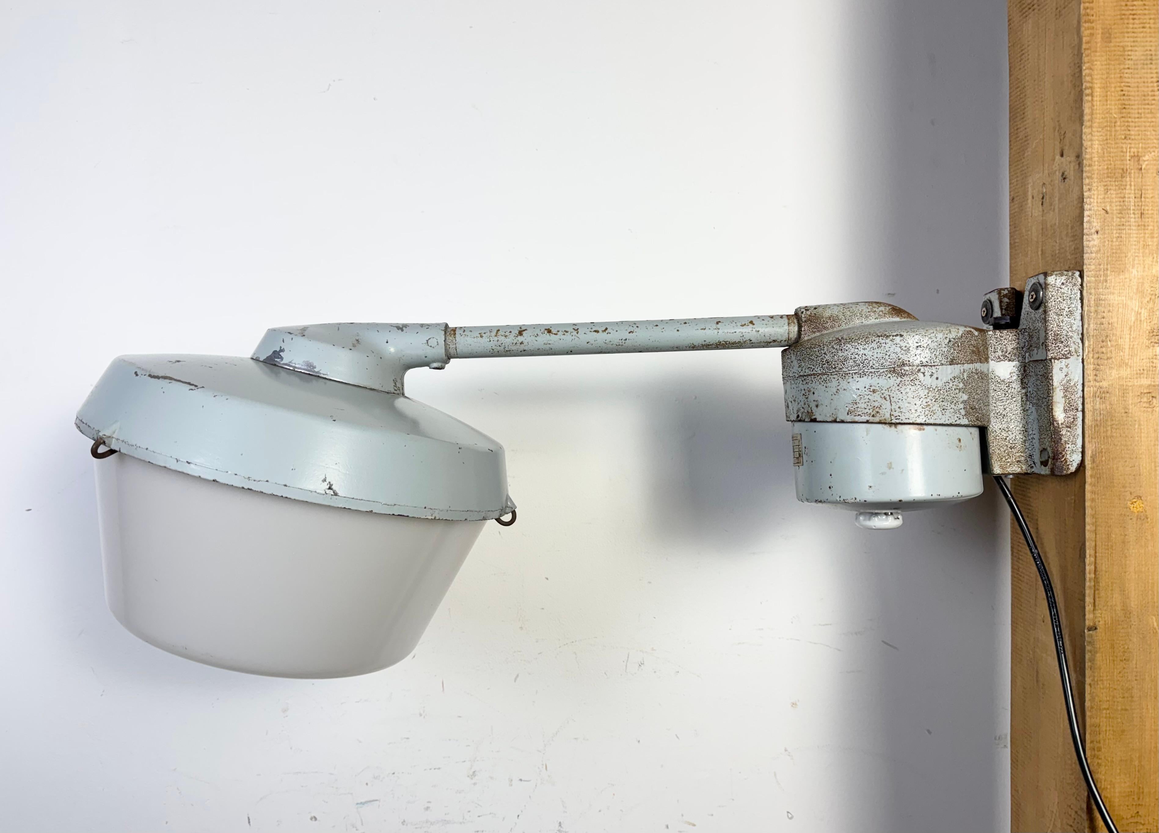 Applique industriale realizzata nell'ex Cecoslovacchia negli anni Settanta. Presenta un braccio e un supporto da parete in ghisa grigia, un paralume in alluminio fuso grigio e un coperchio in vetro lattiginoso in plastica.  La presa originale