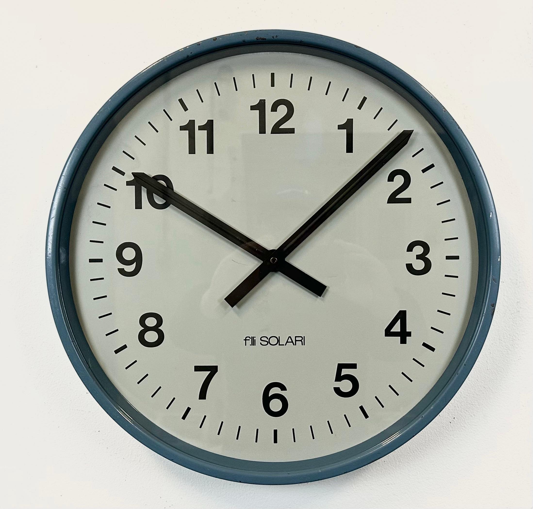 This wall clock was produced by Fratelli Solari Udine in Italy during the 1970s. It features a blue metal frame, an iron dial, an aluminium hands and a clear glass cover. The piece has been converted into a battery-powered clockwork and requires