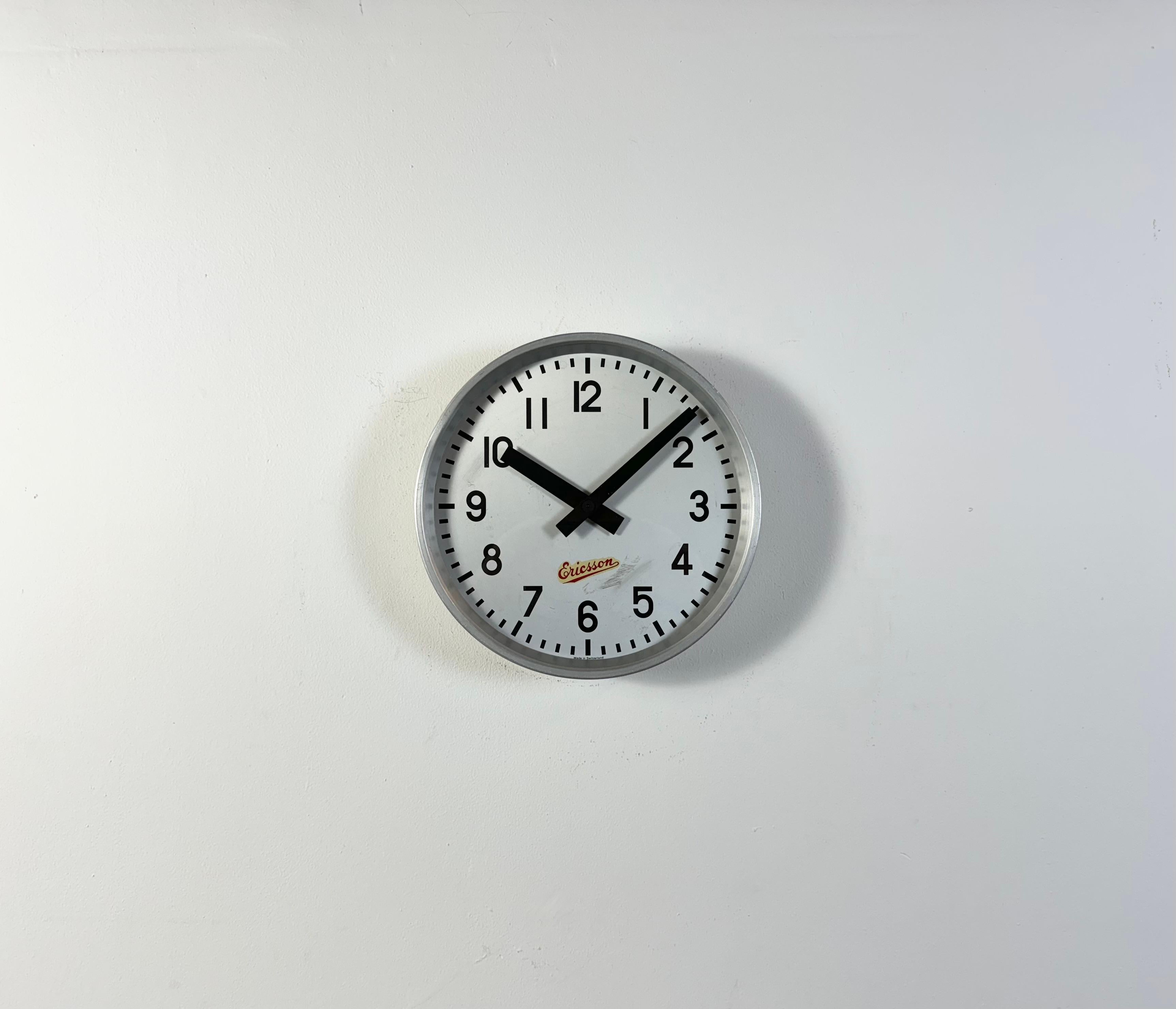Reloj de pared industrial vintage fabricado por Ericsson en Suiza durante la década de 1960. Tiene una estructura de hierro gris, una esfera de metal plateado, agujas de aluminio y una tapa de cristal transparente. La pieza se ha convertido en un