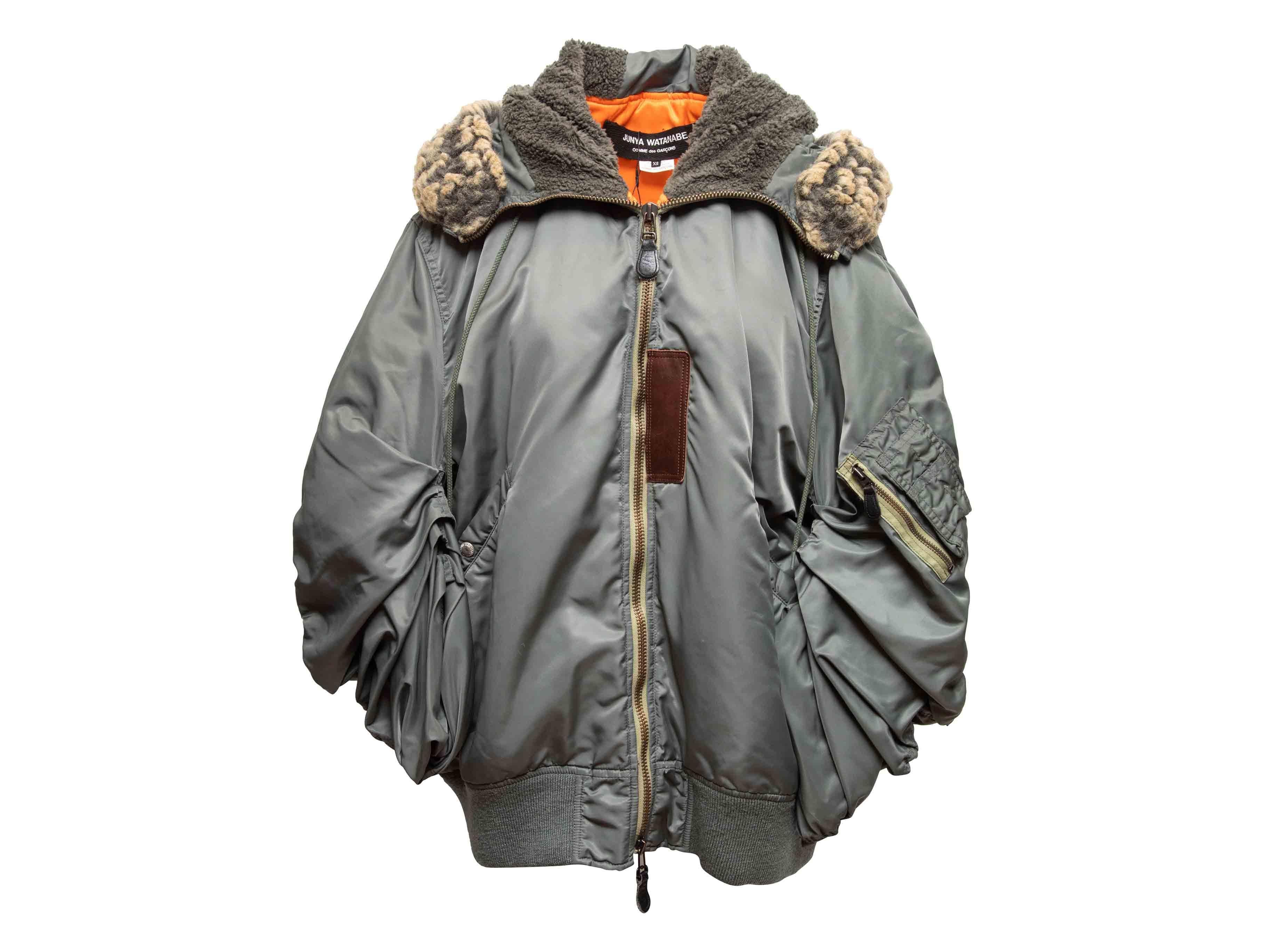 Bomber oversize grigio con cappuccio di Junya Watanabe Comme Des Garcons. Rivestimento in pelliccia sintetica sul cappuccio. Bordi in maglia a coste. Doppie tasche sui fianchi. Chiusura con zip sul davanti. Seno 50