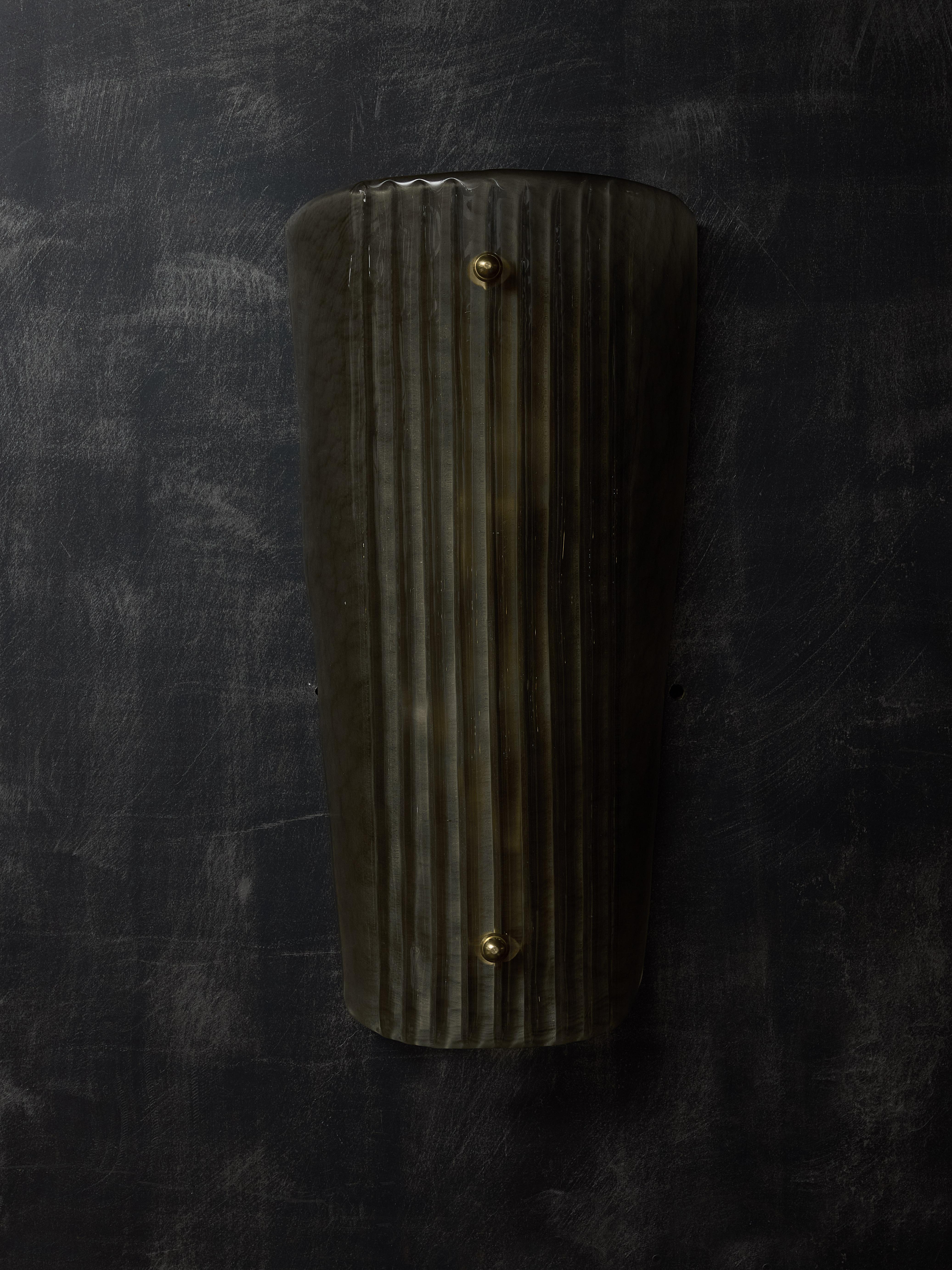 Wandleuchter aus gebogenem Murano-Glas in Form eines Schildes mit vertikalen Rillen, mit kleinen Zierschrauben aus Messing.
Zwei Lichtquellen pro Leuchte