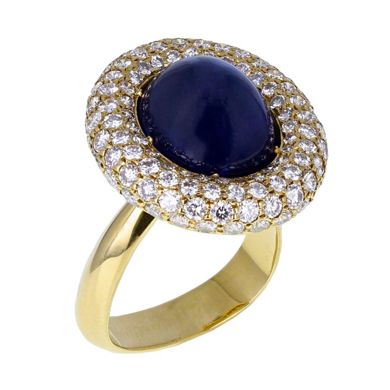 From the famed workshop of Andrew Grima, an exquisite sapphire cocktail ring in 18-carat yellow gold. The central oval-shaped sugarloaf sapphire cabochon of approximately 14.00 carats is nestled in a domed pillow of bright and lively pavé set