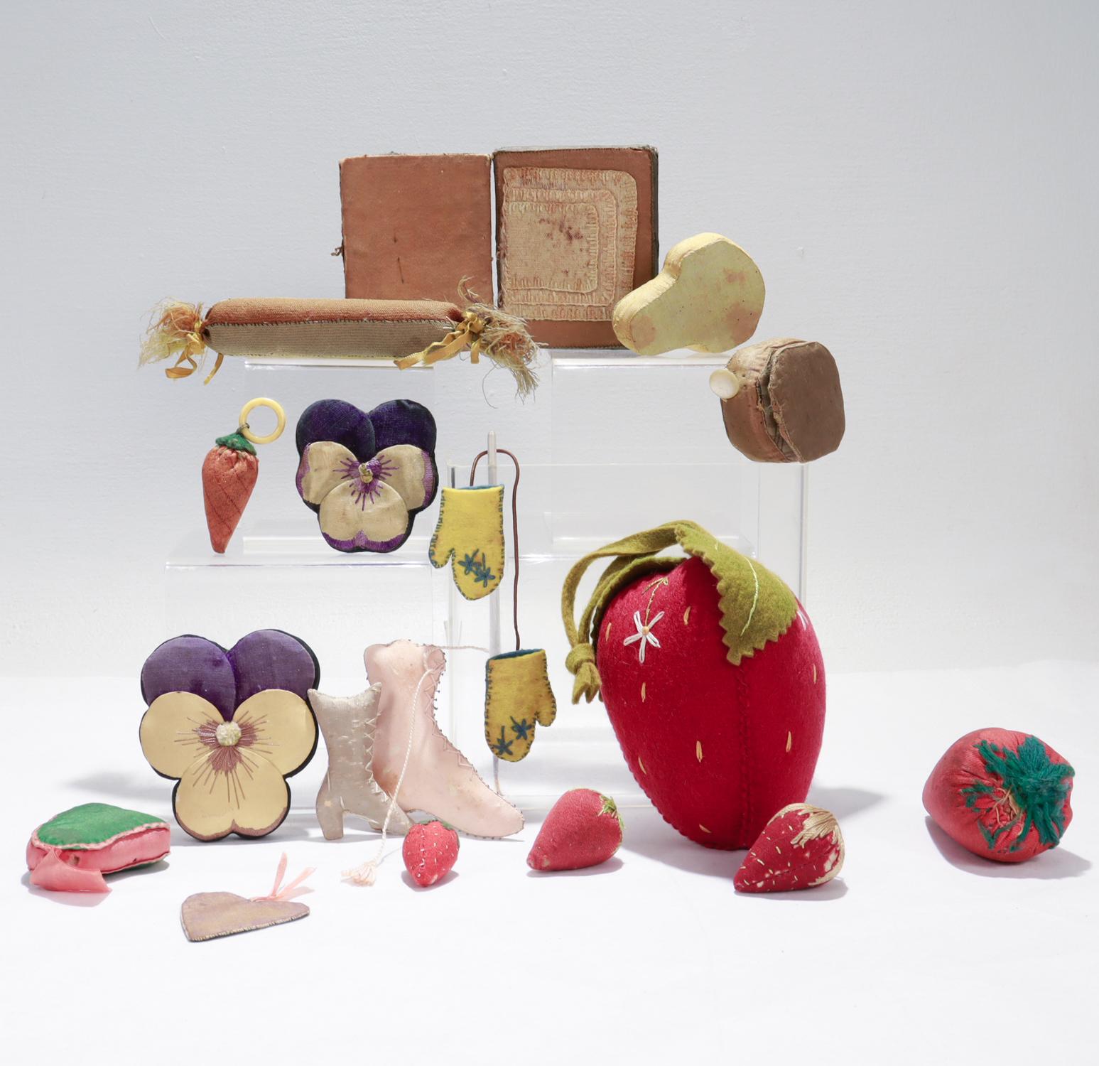 An incredible collection of 17 Folk Art pin cushions.

The group comprising pansy flowers, strawberries, boots, mittens, a rolled cushion, 2 books, a pear form, a bell form, a carrot, and a heart.

In varying materials including silks, wools, and