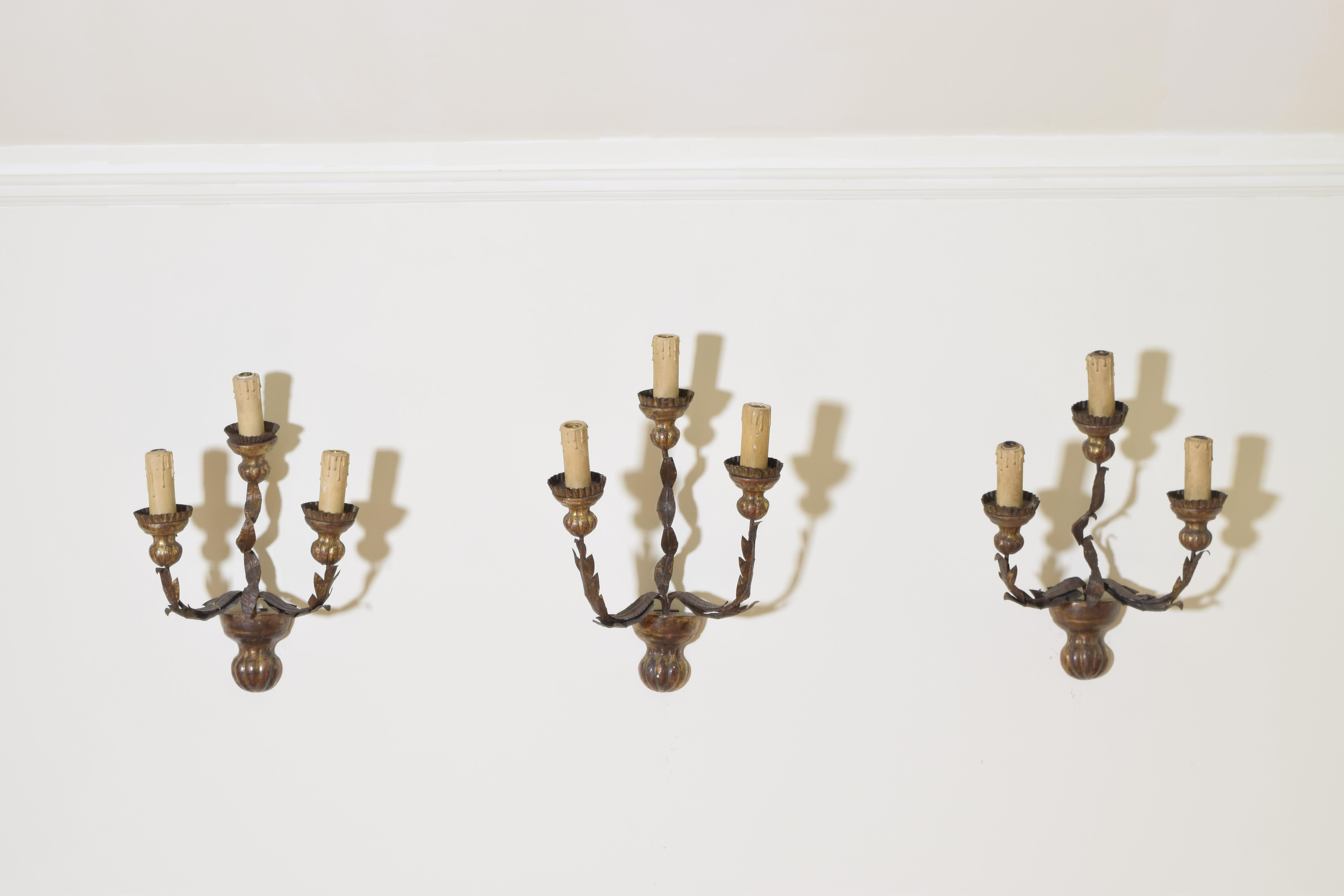 Each sconce having a campana form carved giltwood base issuing three iron arms with applied formerly gilt metal leaves, the carved giltwood bobeches of similar form as the bases and having reticulated metal drip pans, offered as a set of 6 or as
