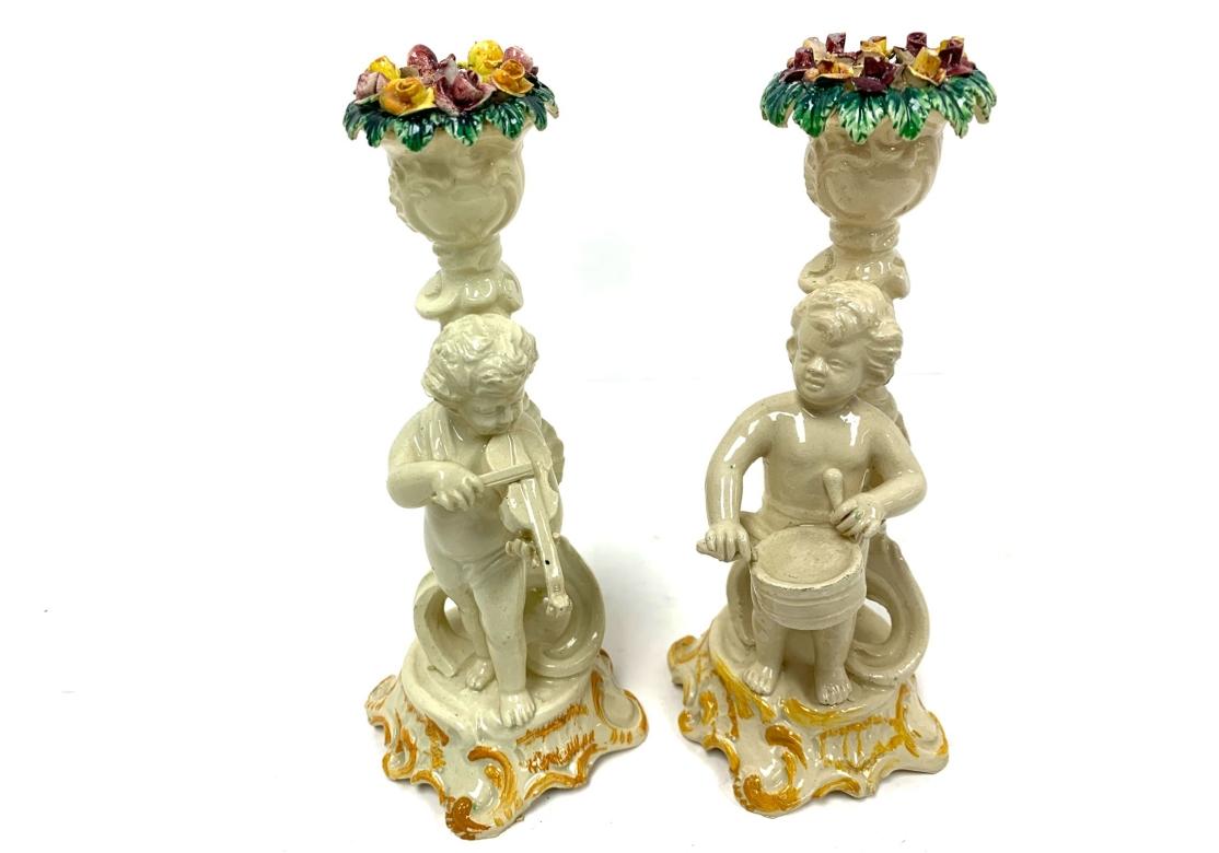 Group of four Italian pottery candlesticks with four putti with musical instruments. The putti are standing in front of a seashell form column and surmounted by a della robbia style urn. The putti are standing on a hand painted plinth base, each
