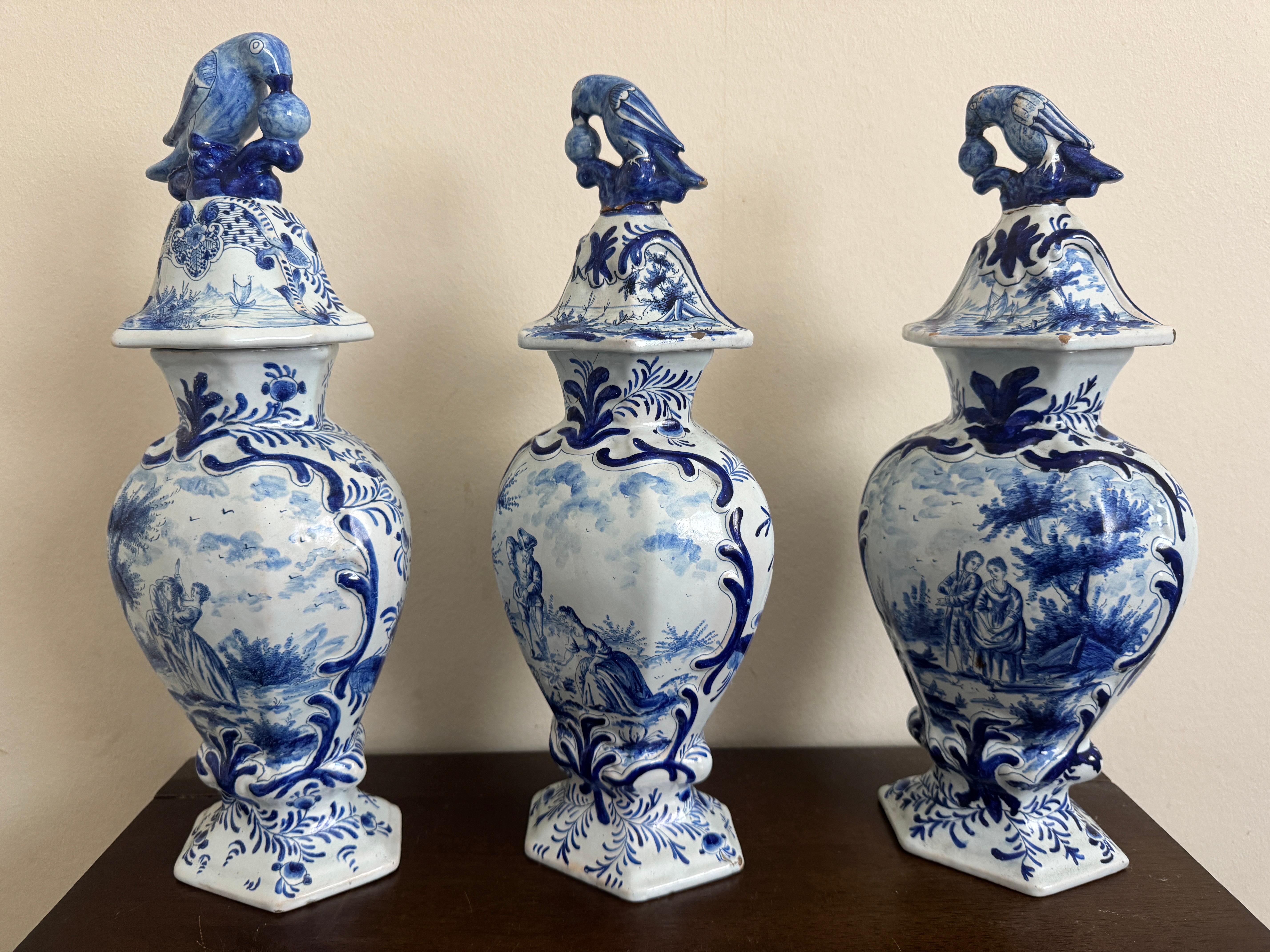 Group of Three 18th Century Delft Jars and Lids, blue and white, each painted with figural vignettes and bearing bird & ball finialled covers., with maker's marks possibly indicating a link to Johannes Harlees at De Porceleyne Fle.

.