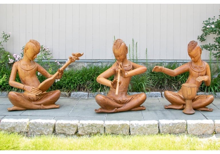 A set of three large heavy carved teak musician figures. All are seated in elegant dance- like poses and wear local style loincloths, neck torques and anklets. One plays a stringed instrument, one a flute, and the third a drum. Each separately made