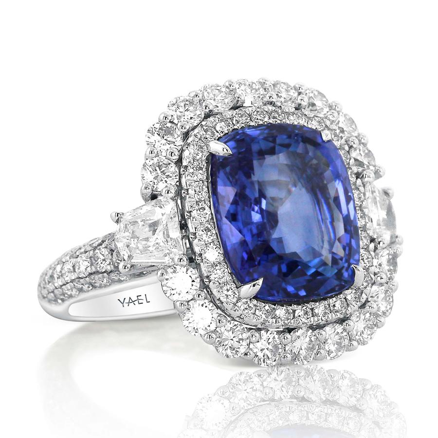 A limpid blue sapphire within a diamond halo is the central feature of this graceful ring. They lay within a second halo divided by shield-shaped diamonds at either shoulder, whose hard lines accent the softer curves of the piece. A diamond-set