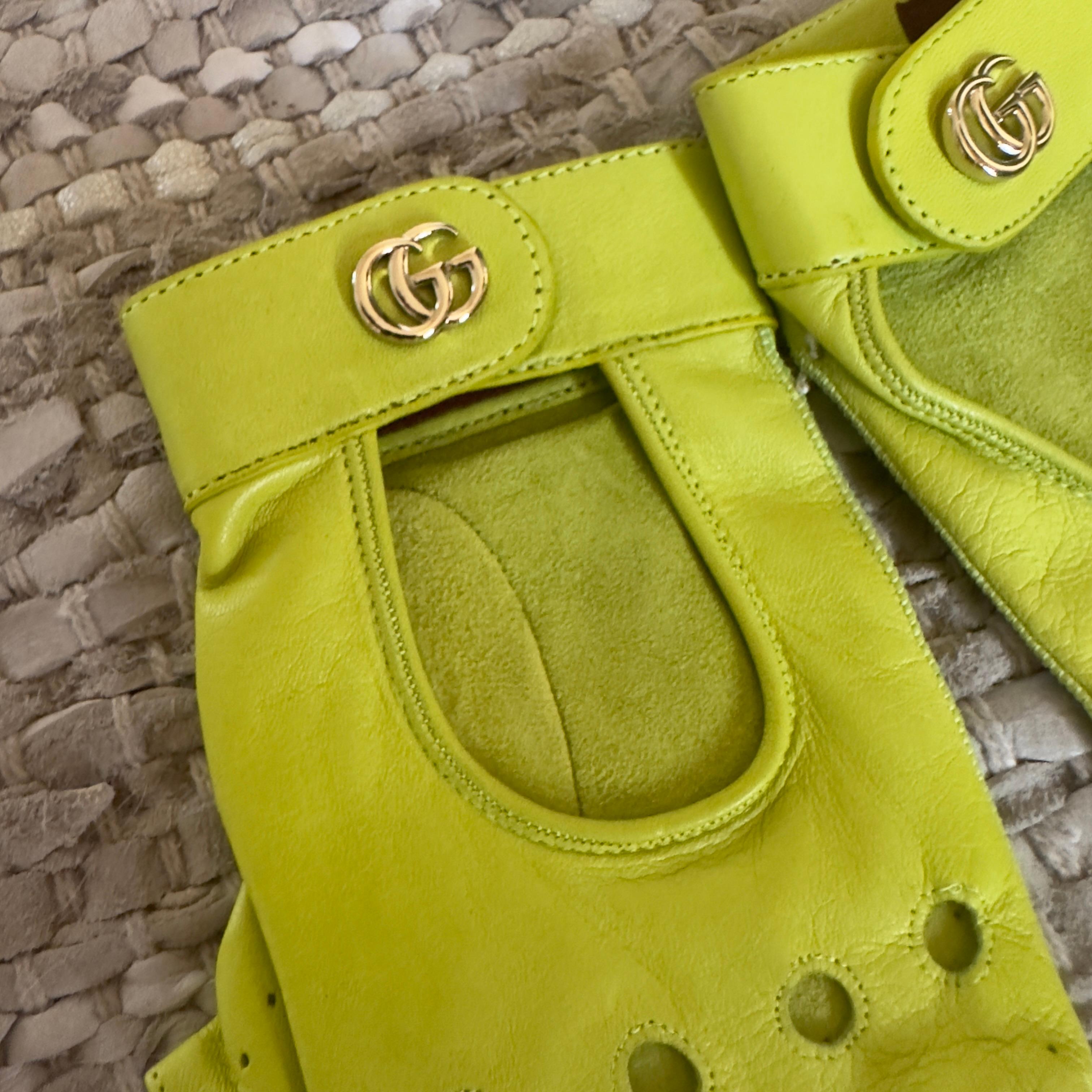 Guanti da guida Gucci in pelle liscia gialla limone, parte della collezione attuale del brand. Modello senza dita con taglio ergonomico, traforature decorative e iconico logo GG in metallo dorato sul cinturino. Un accessorio dal design audace e