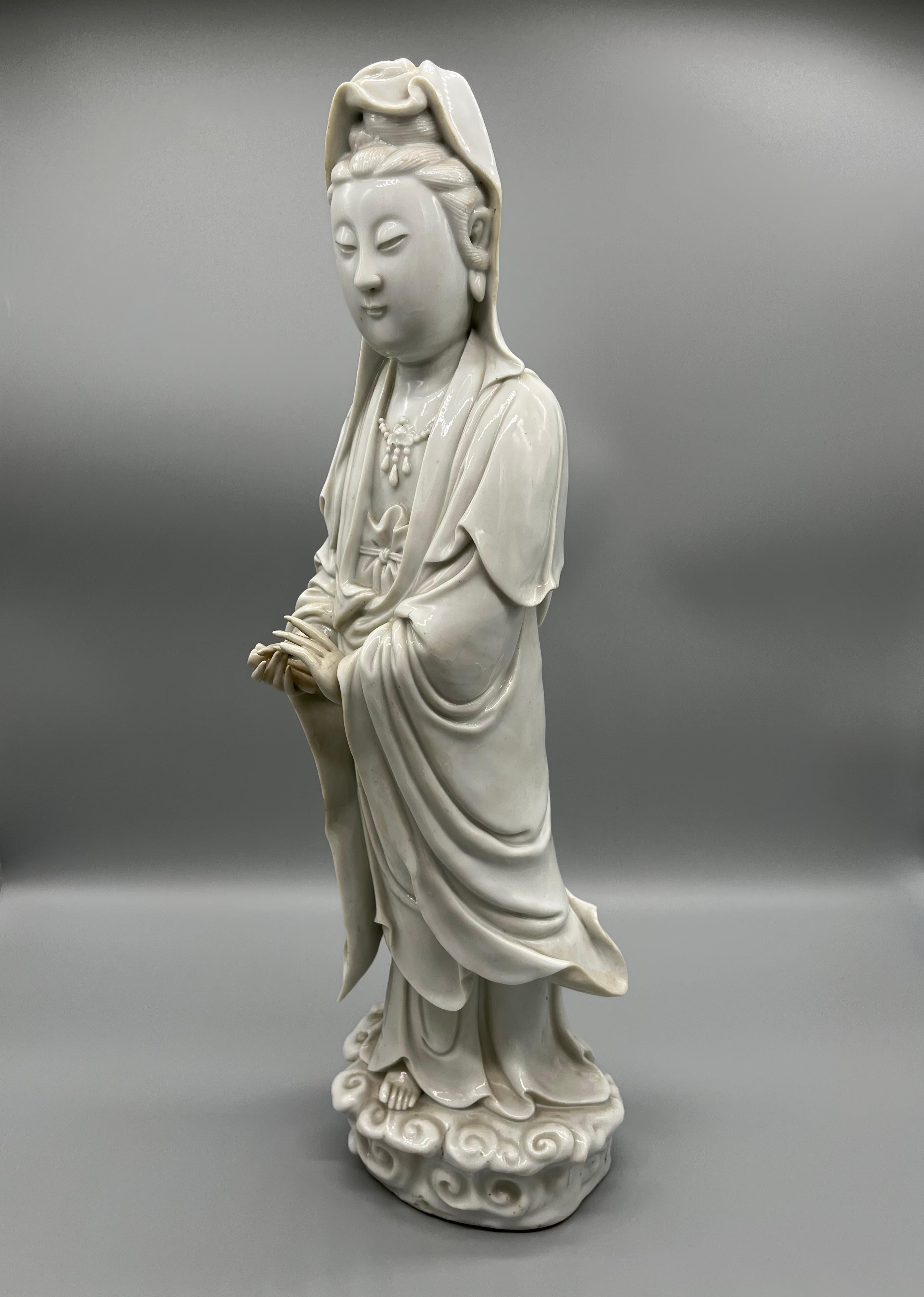 A Blanc-de Chine Figure of Guanyin (goddess of mercy) standing on a pedestal of waves, the arms lifted to the waist, dressed in loose fitting robes opening at the chest to reveal a necklace, applied with an ivory glaze. China antique.